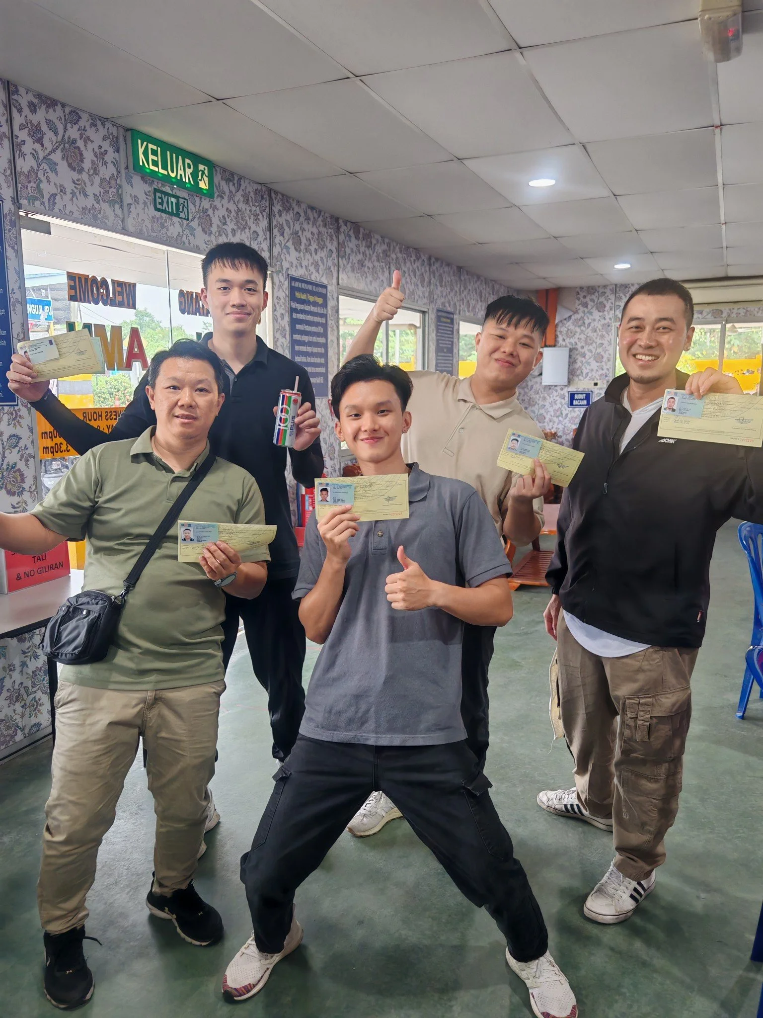 Six men posing indoors, holding up paperwork, some smiling and making thumbs-up gestures. The room has floral wallpaper and windows showing outdoor scenery.
