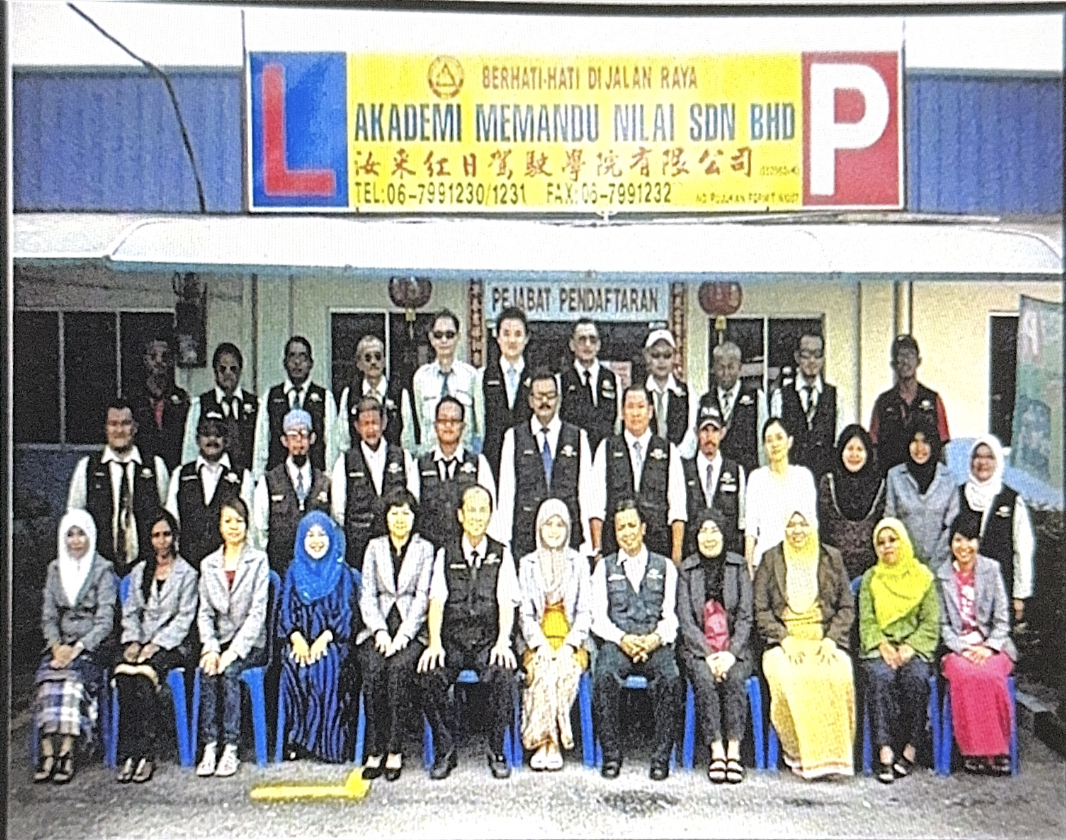 Group of people posing outside a building with a sign that reads 'Akademi Memandu Nilai SDN BHD' and a large parking sign with an 'L' and 'P' for learners and parking.