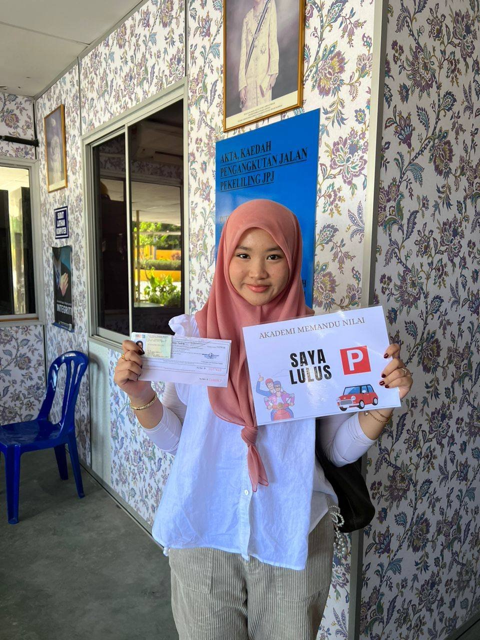 Young woman wearing a pink hijab and white shirt holding a driving test pass certificate and a sign that says "Saya Lulus P" with illustration of a red car, smiling inside an indoor setting with floral wallpaper.