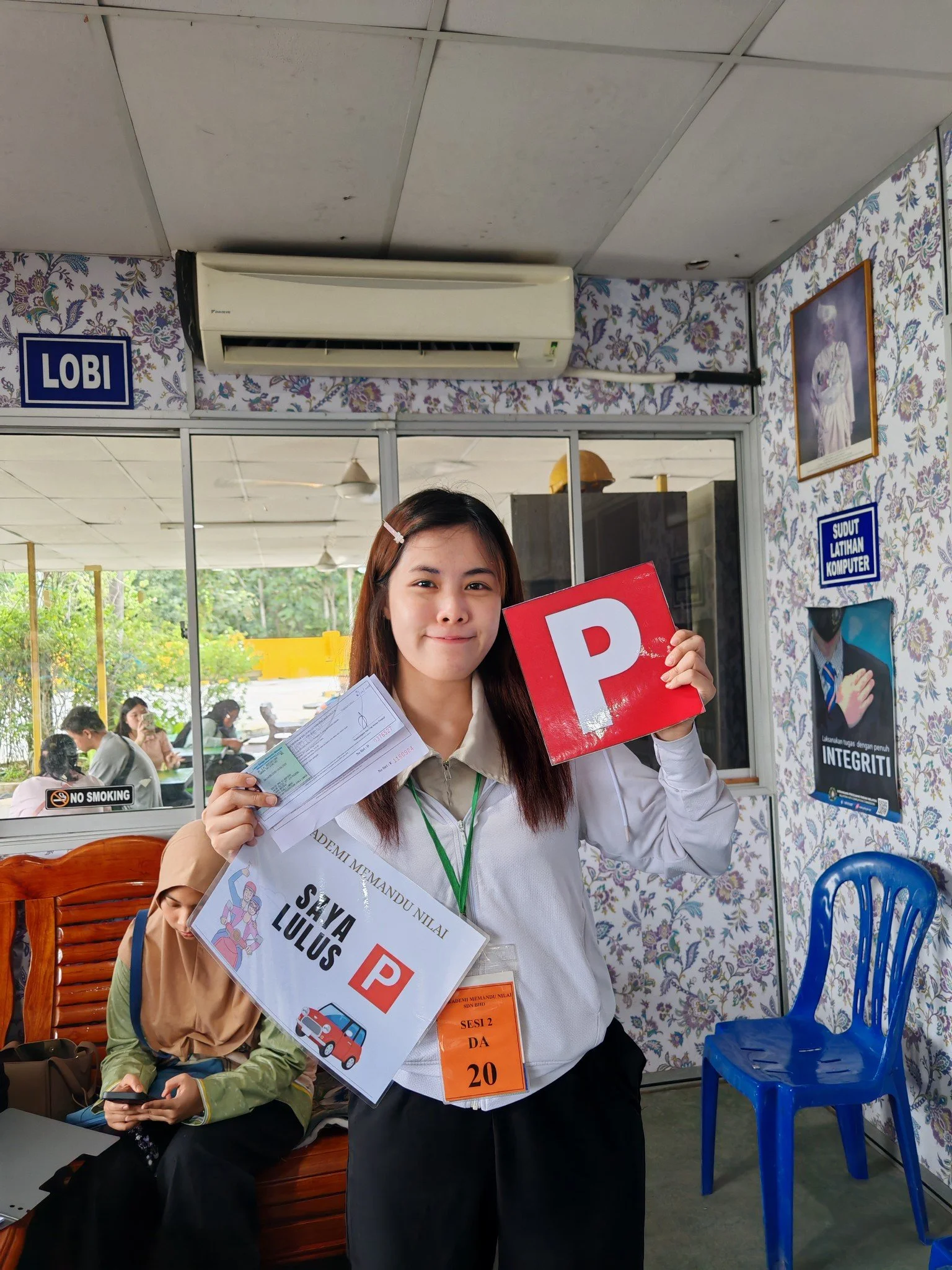 Young woman at a driving school, holding up a 'P' sign for 'probation' and her driving test pass certificate. She wears a white shirt with a name tag and smiles at the camera. Background shows other people inside the building and outdoor greenery.