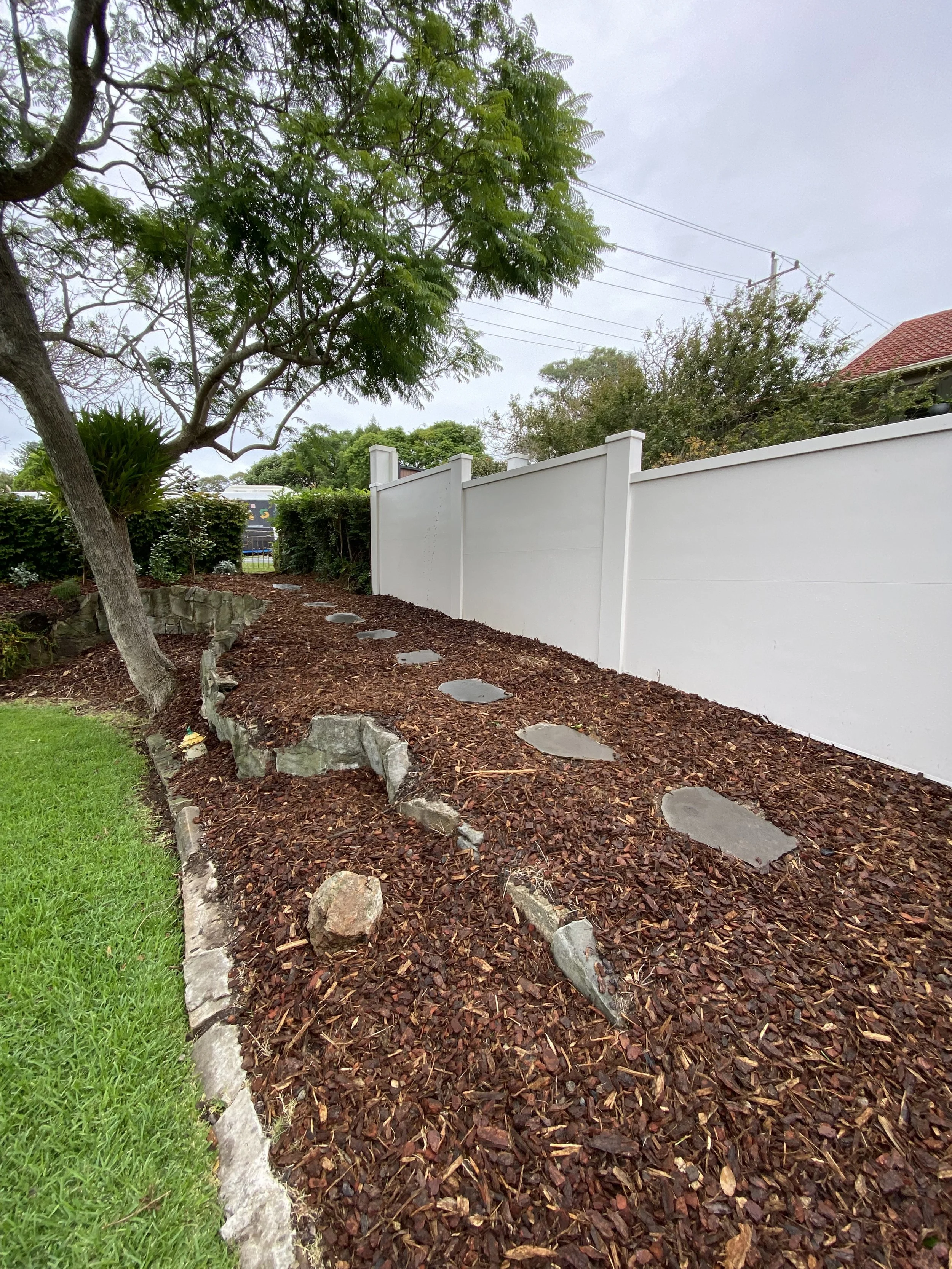Pathway with stepping stones and mulch next to white fence in garden setting.