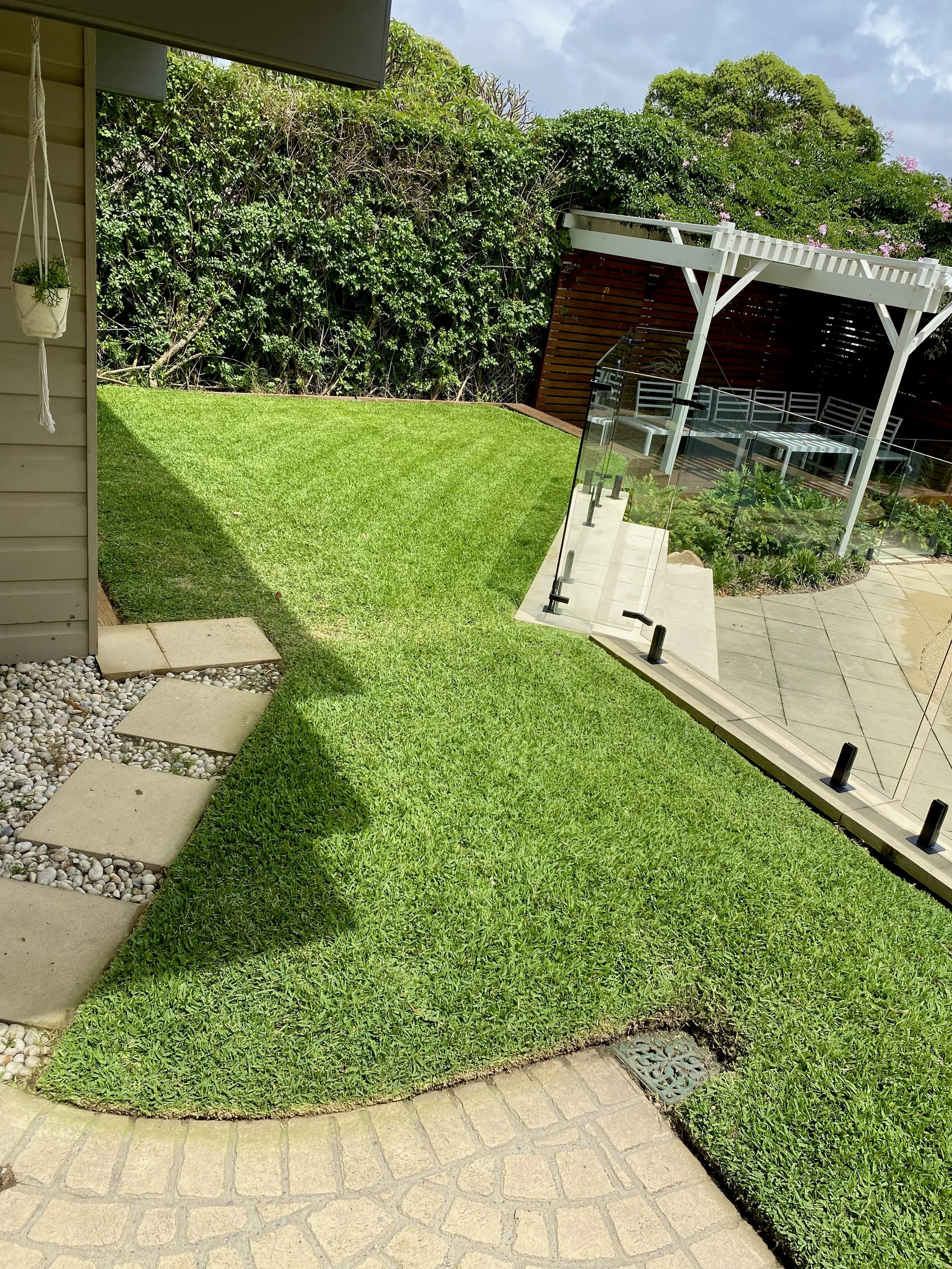 Backyard with a well-maintained lawn, a wooden pergola with seating, and a glass barrier next to a paved area. There's a pebble pathway and plants along a wooden fence with greenery.