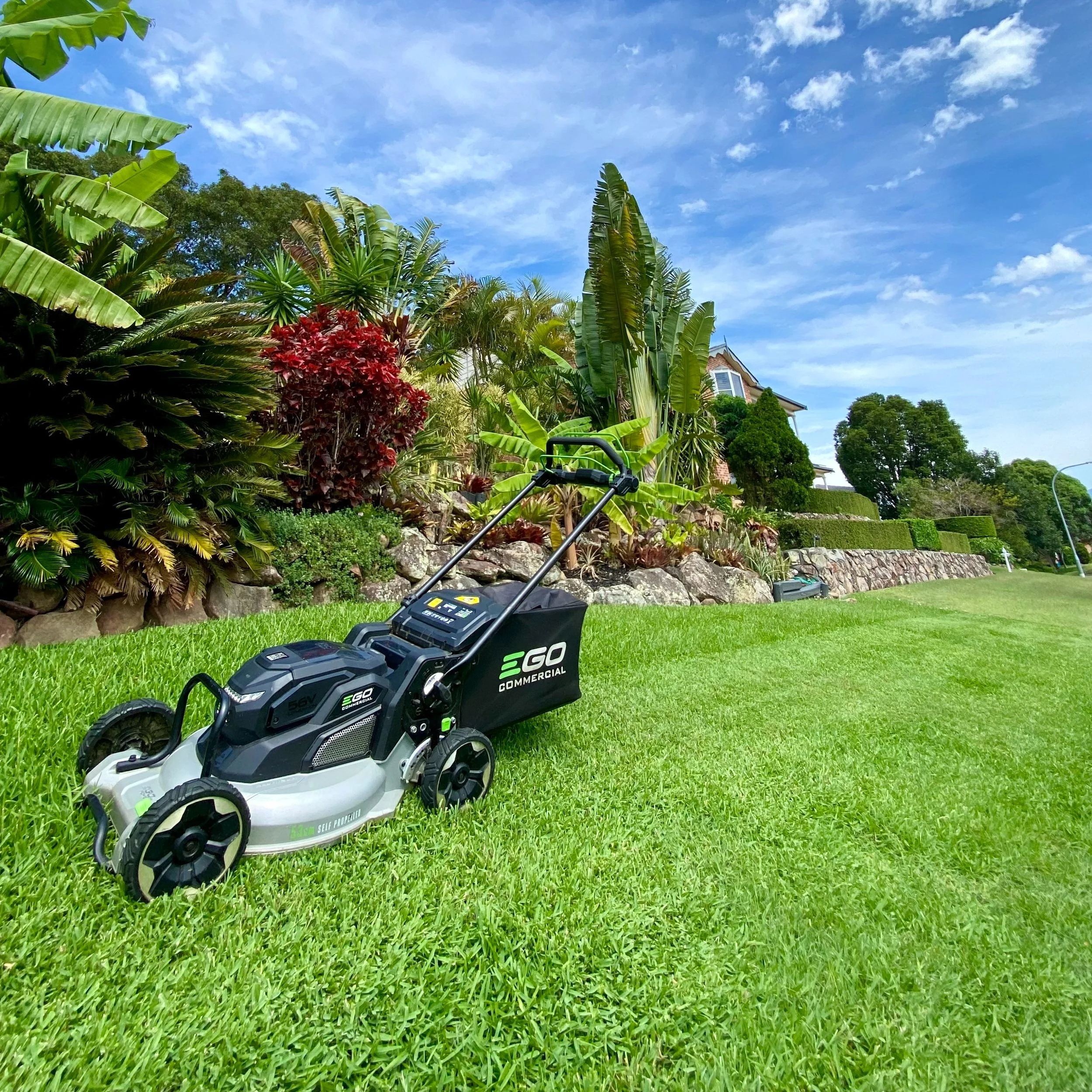 Electric lawn mower on green grass with landscaped garden and trees in the background.