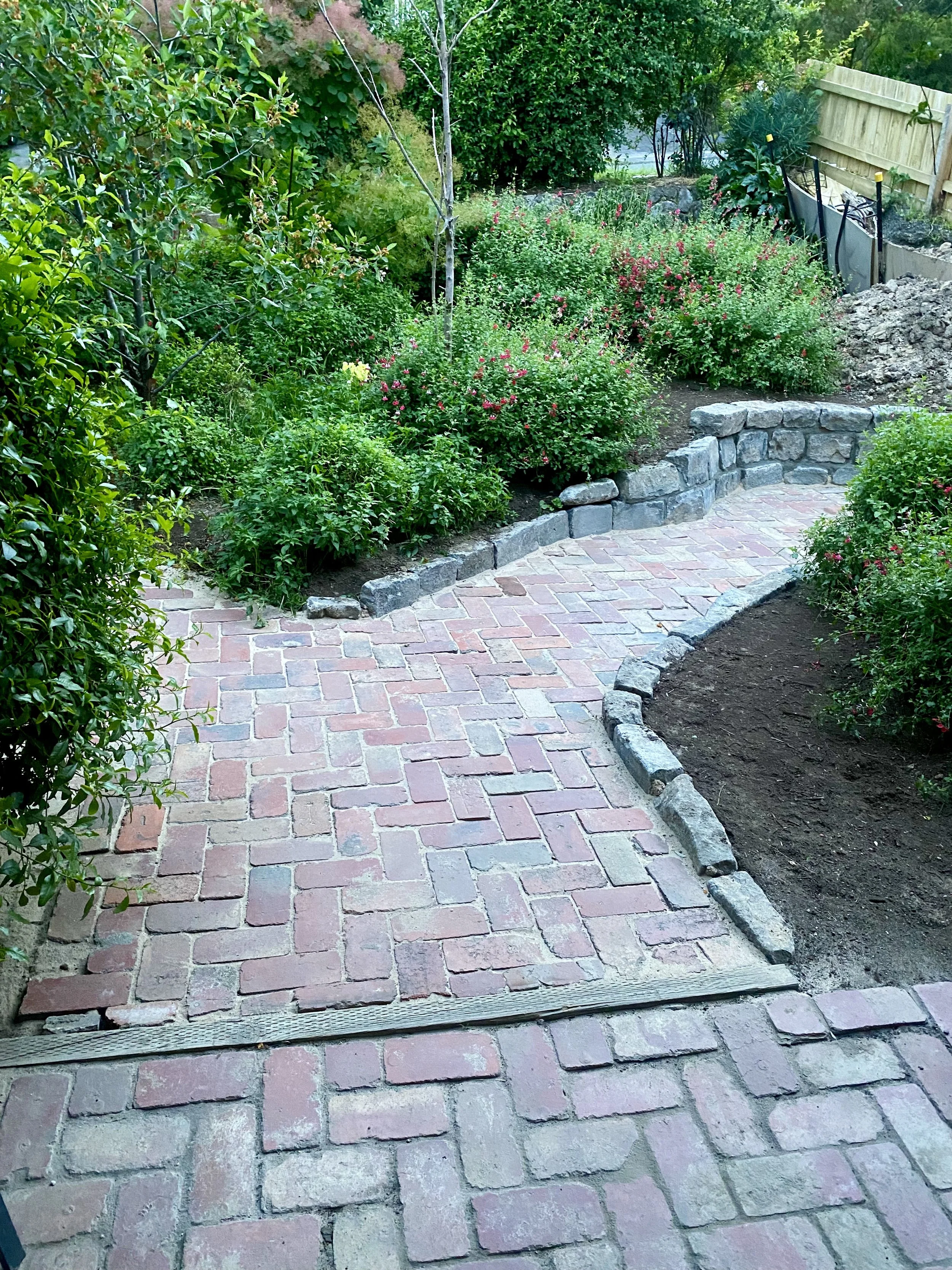 Brick pathway surrounded by lush green bushes and plants, with a low stone wall and a wooden fence in the background.