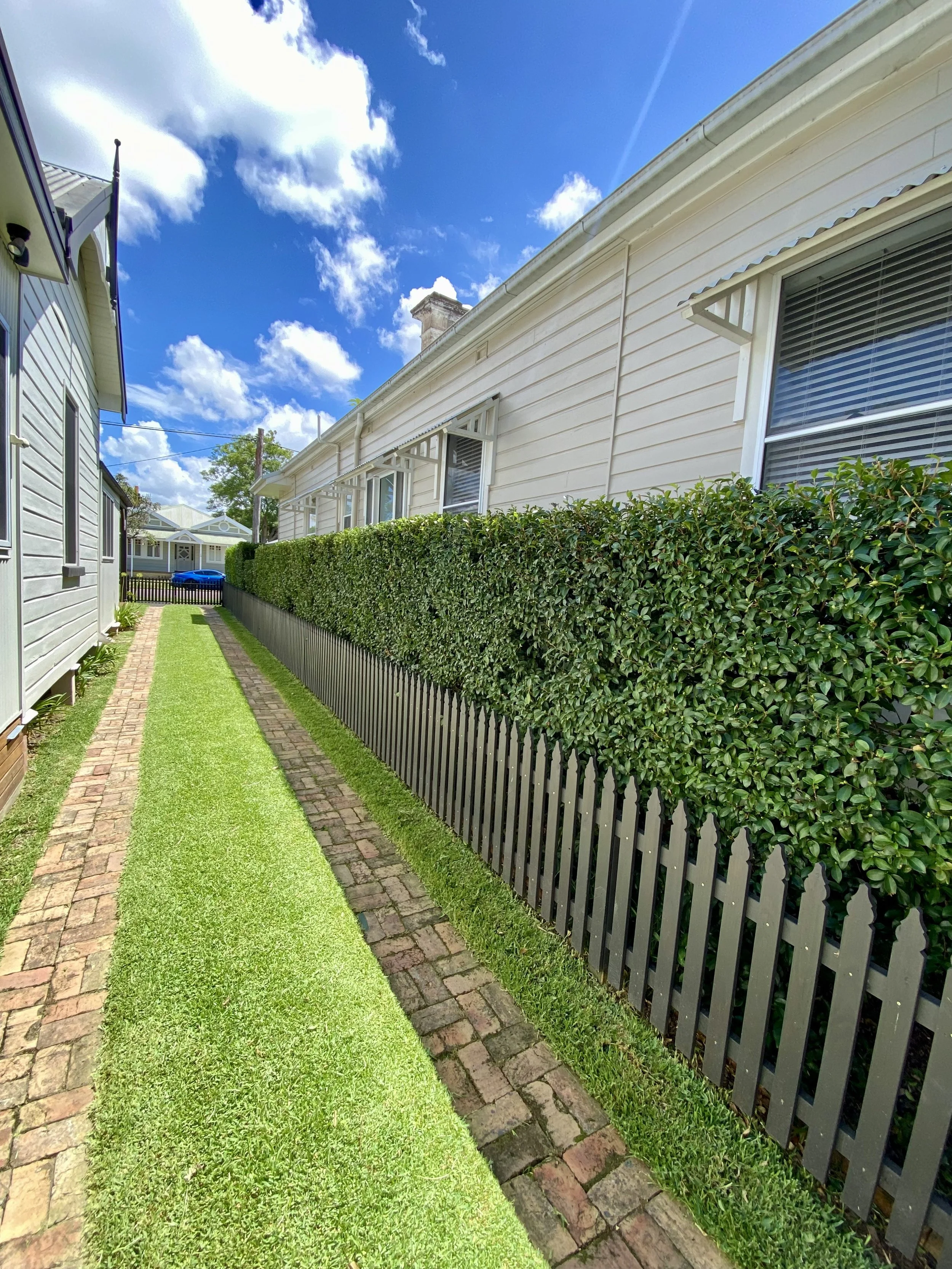 A narrow alley between two houses with a brick and grass path, bordered by a wooden picket fence and a hedge on one side. The skies are clear and sunny with some clouds.