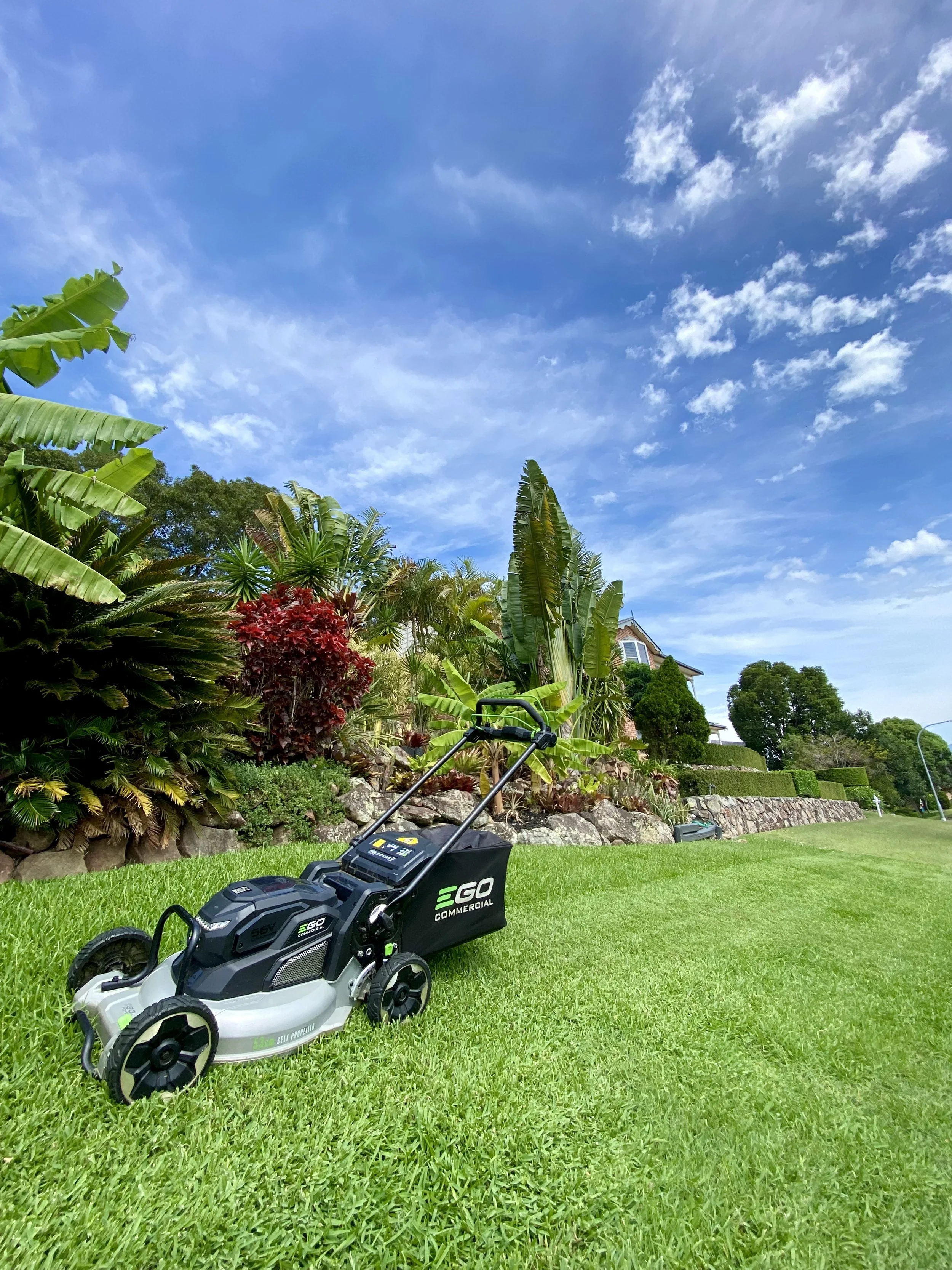Electric lawn mower on neatly cut grass, surrounded by tropical plants and beneath a blue sky with scattered clouds.