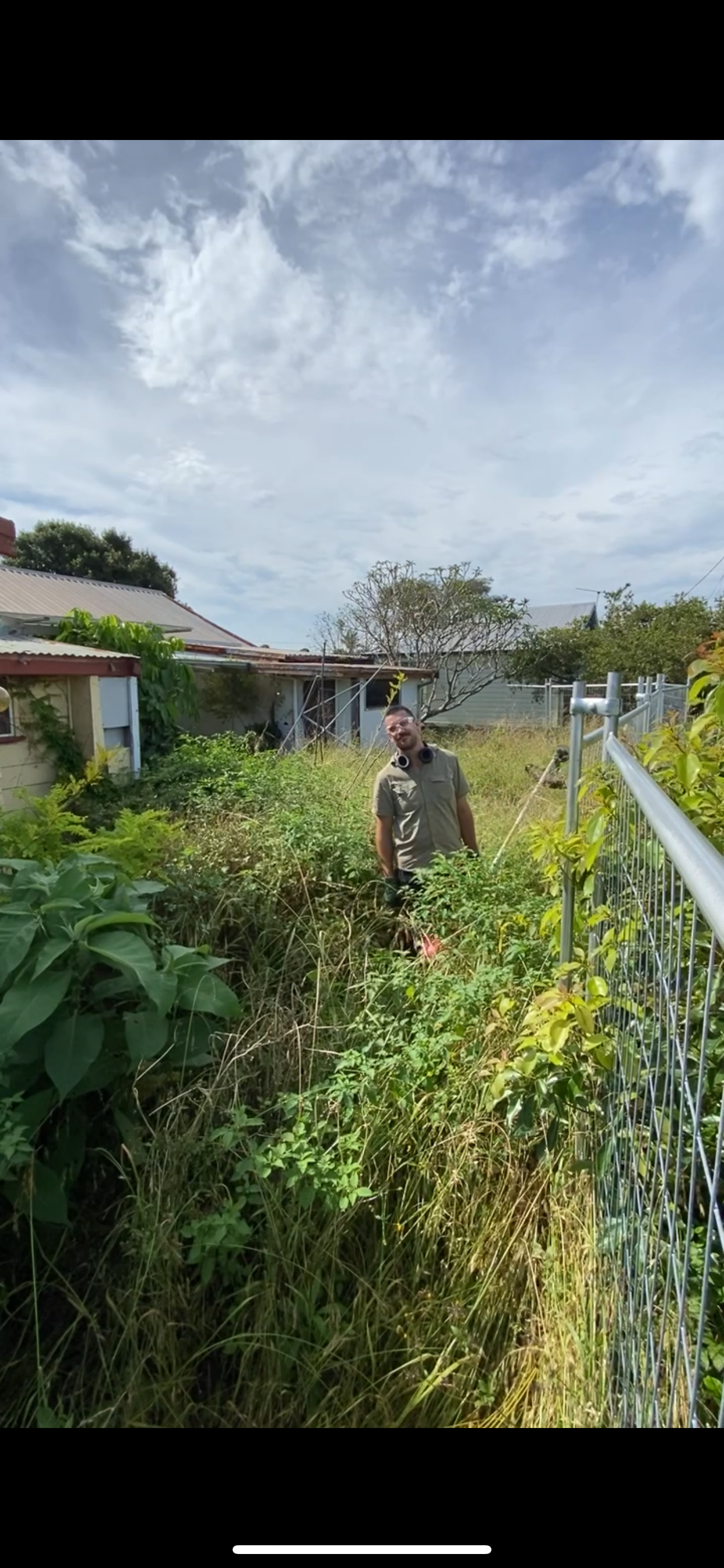 A person stands in an overgrown garden with tall grass and plants, beside a fenced area and an old house with a corrugated roof. The sky is partly cloudy.