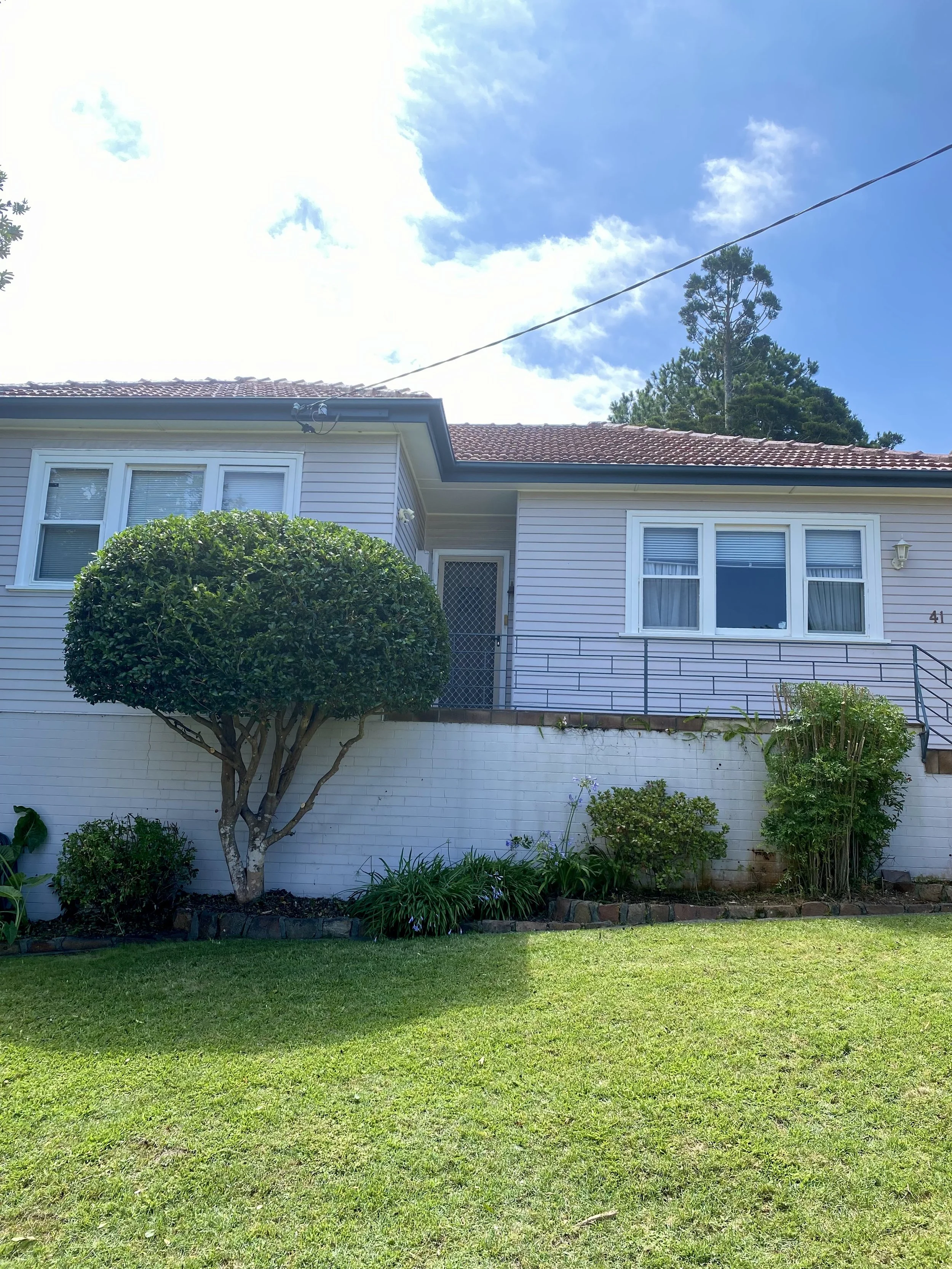 Single-story house with a tiled roof, white siding, and a manicured lawn. There is a trimmed tree and various plants in the front yard.