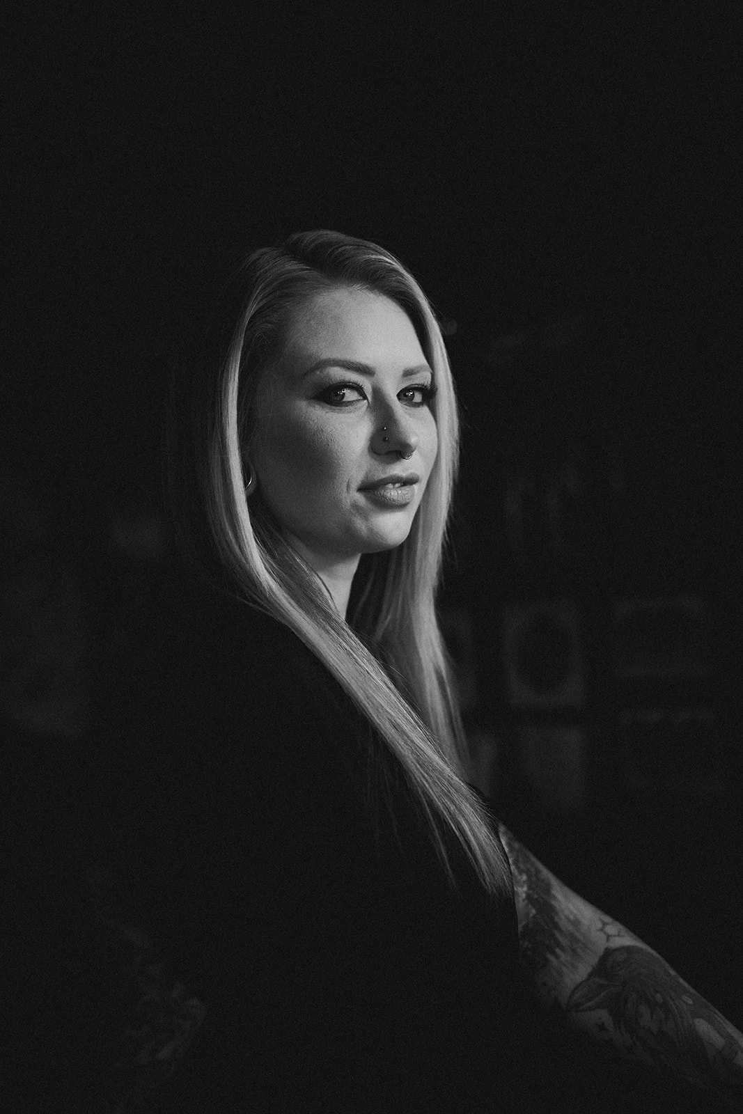 A black and white portrait of a woman with long blonde hair, tattoos on her arm, and facial piercings, looking at the camera with a slight smile.