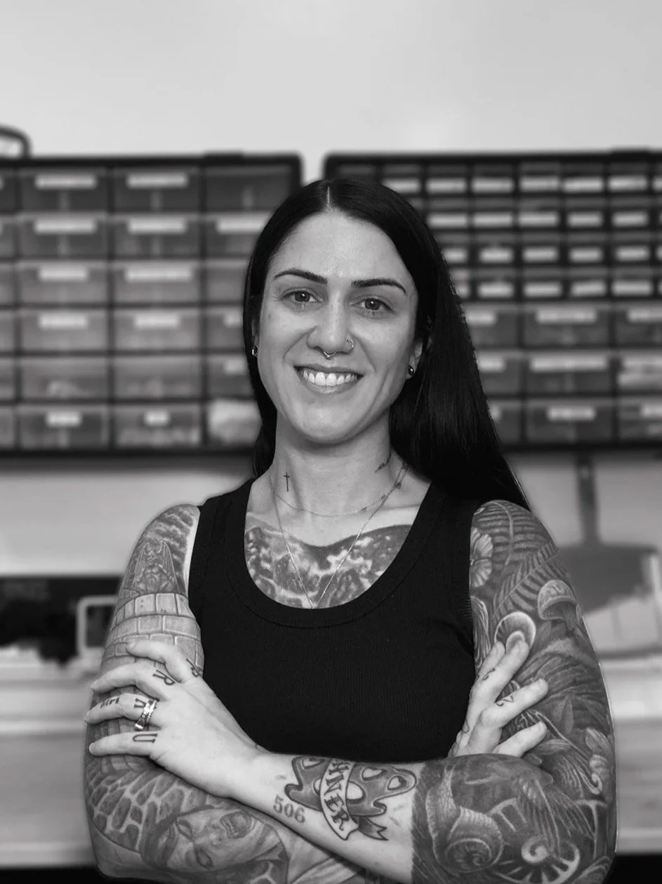 A woman with dark hair and numerous tattoos, smiling with arms crossed, standing in front of a background of small storage drawers.