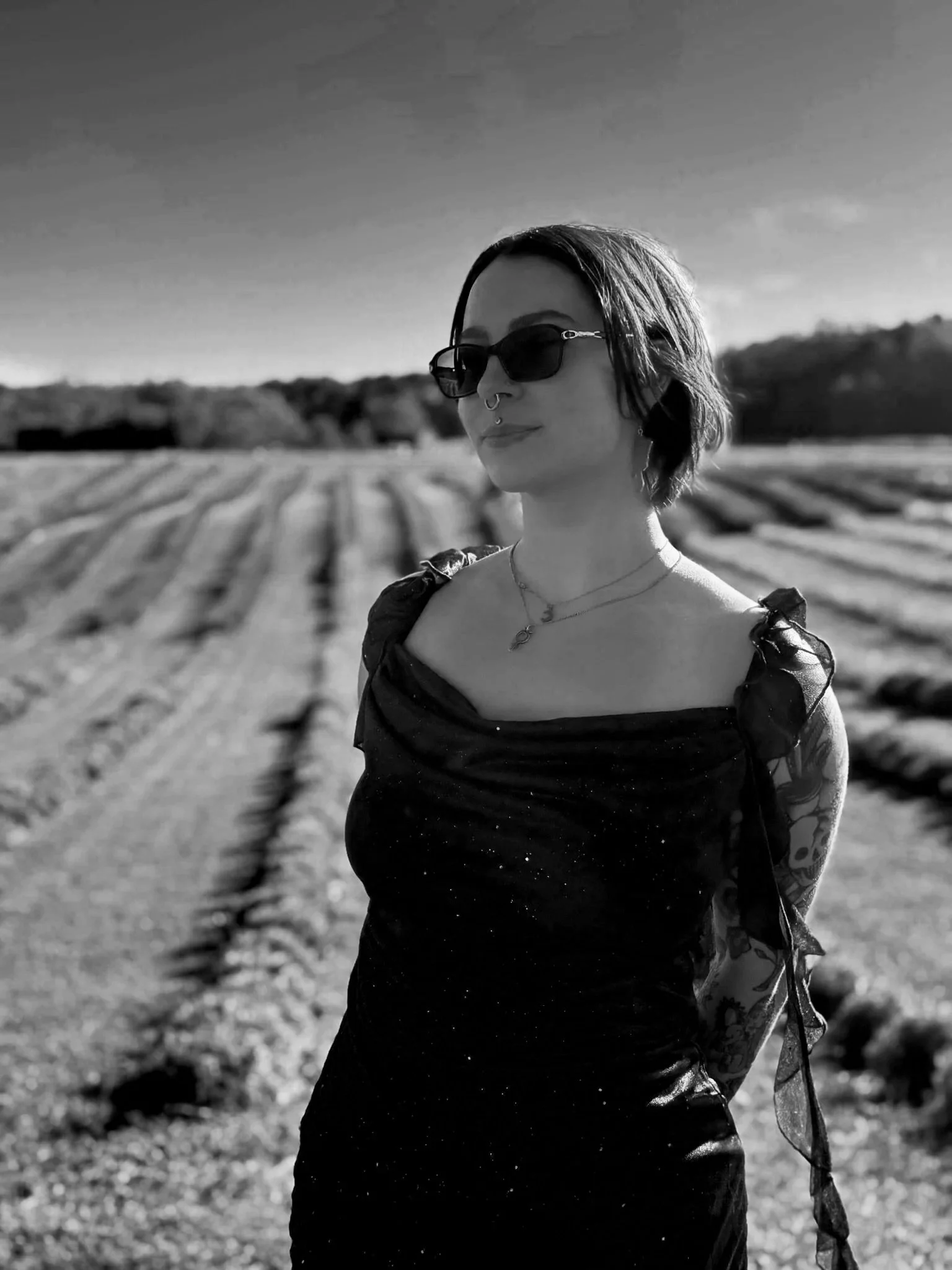 Black and white photo of a woman wearing sunglasses and a black dress, standing outdoors with rows of farmland in the background.