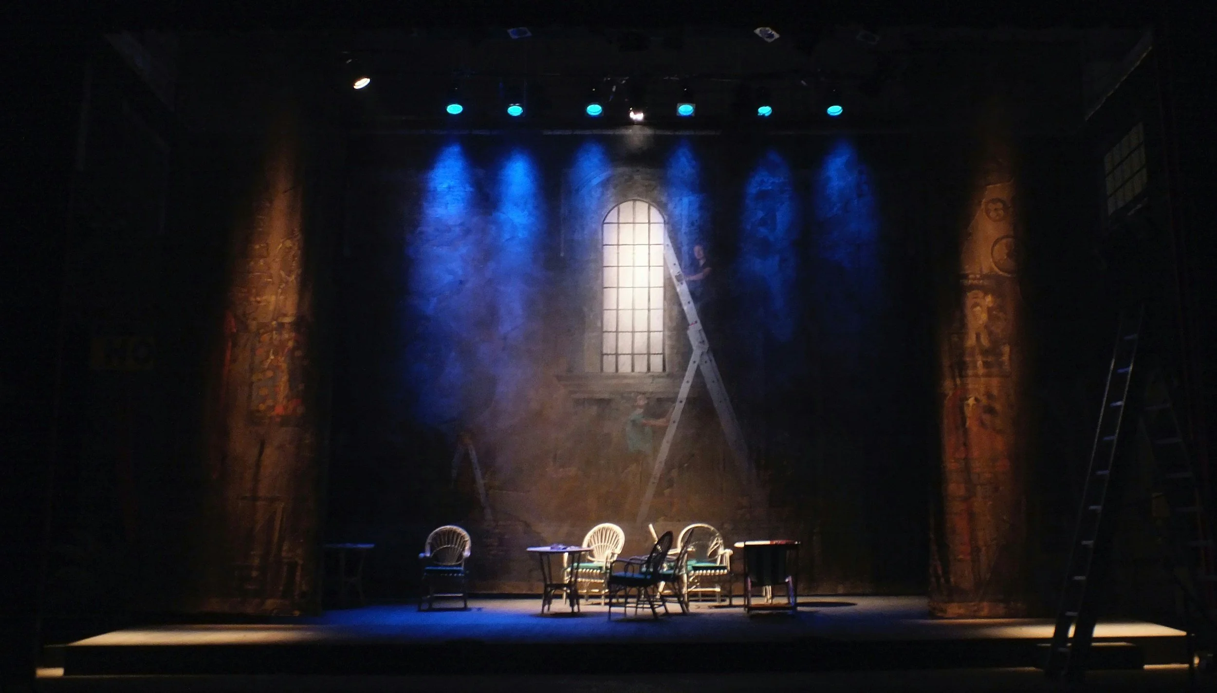 Theater stage set with a window backdrop, blue and warm lighting, four chairs, two small tables, and ladders on both sides.