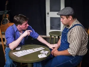 Two men sitting at a table playing cards, with one pointing and the other looking serious, in a room with chairs and a door in the background.