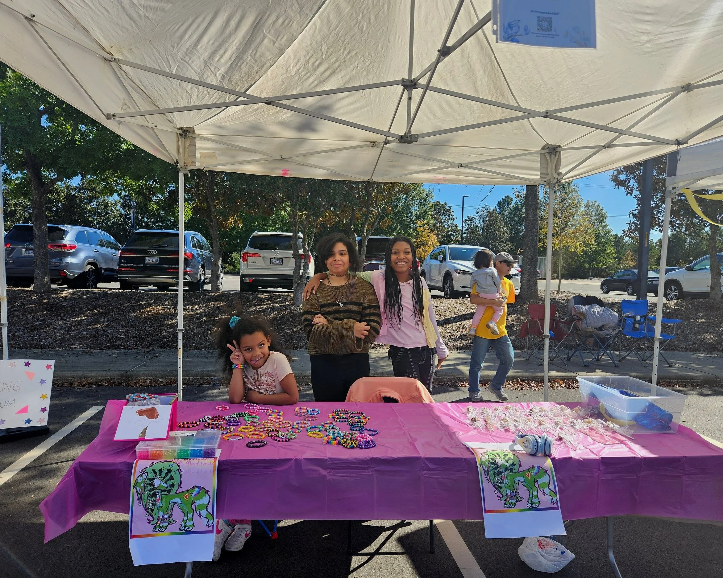 Outdoor craft stall with people, colorful beaded bracelets, and a pink tablecloth under a canopy tent.