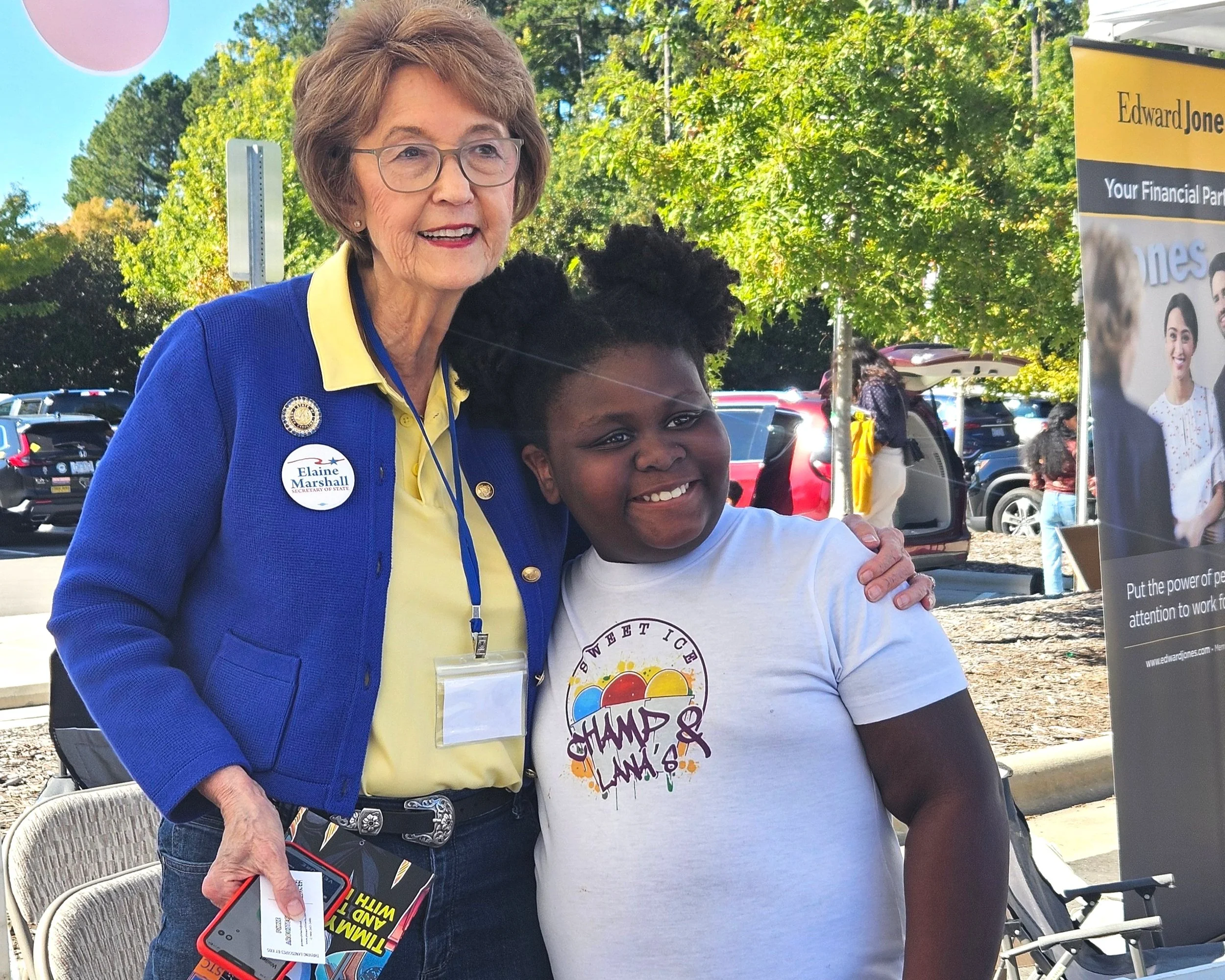Two people smiling and posing for a photo outdoors, surrounded by trees and cars. One person is wearing a blue jacket with a button, and the other is wearing a white T-shirt with colorful text.