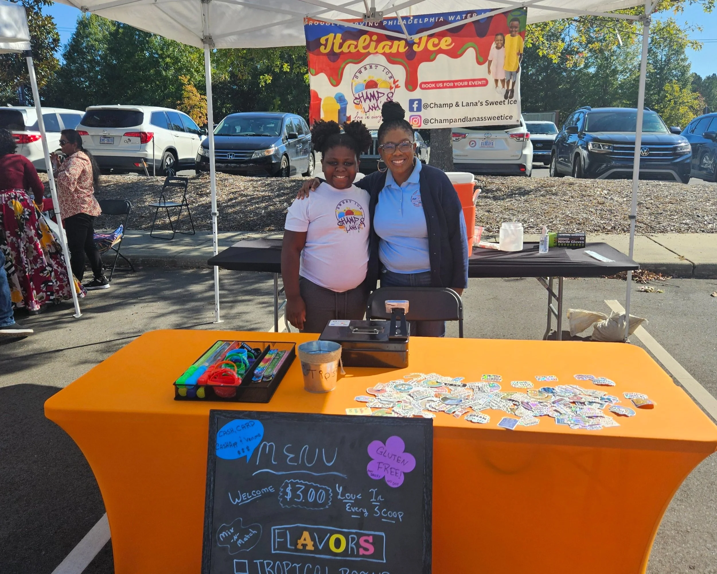 Two people smiling at an Italian ice stand under a tent with a bright orange table and colorful menu sign.