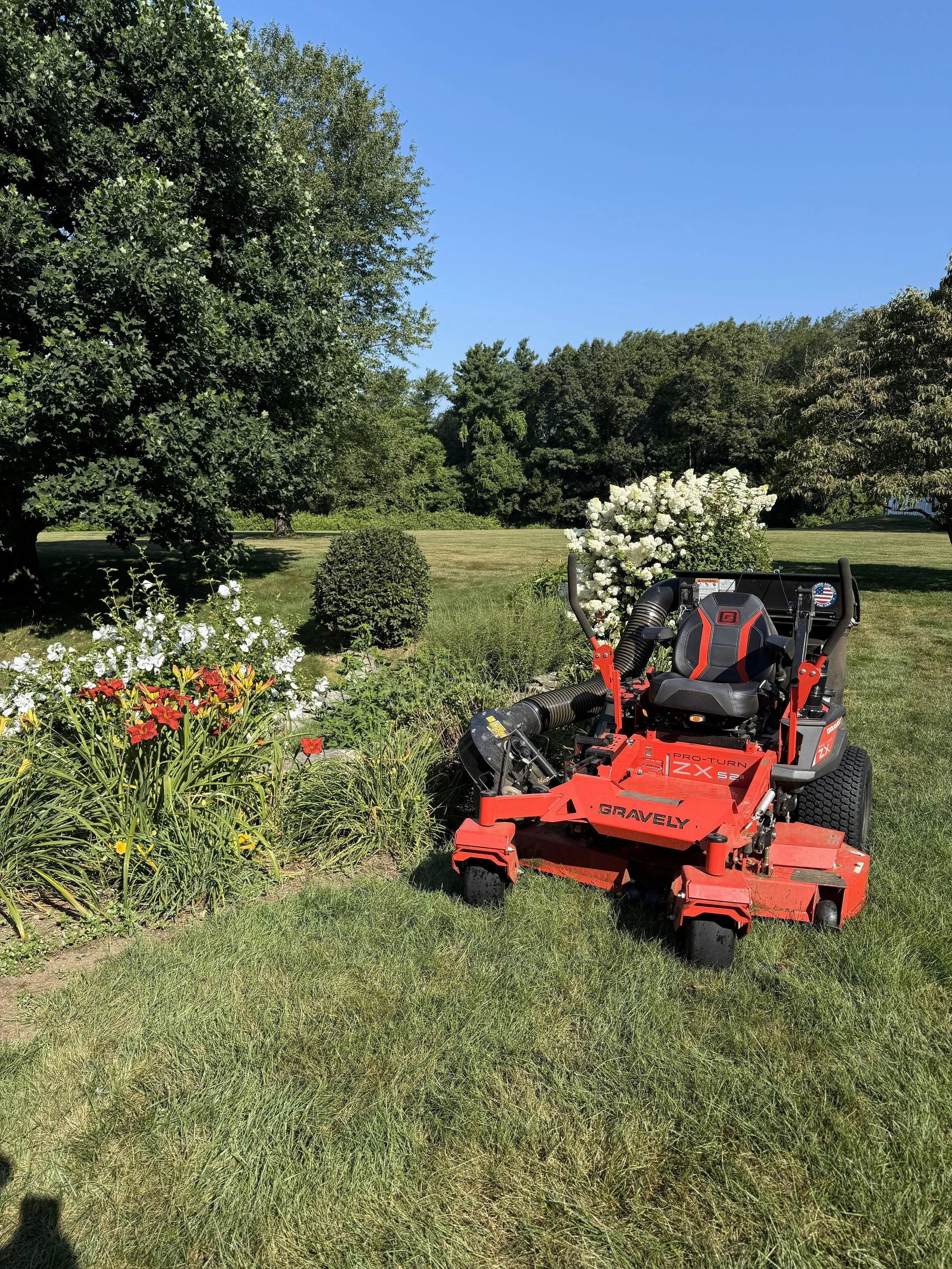 Red lawn mower parked on a grassy lawn next to flowerbed with white and red flowers, trees in the background, clear blue sky.