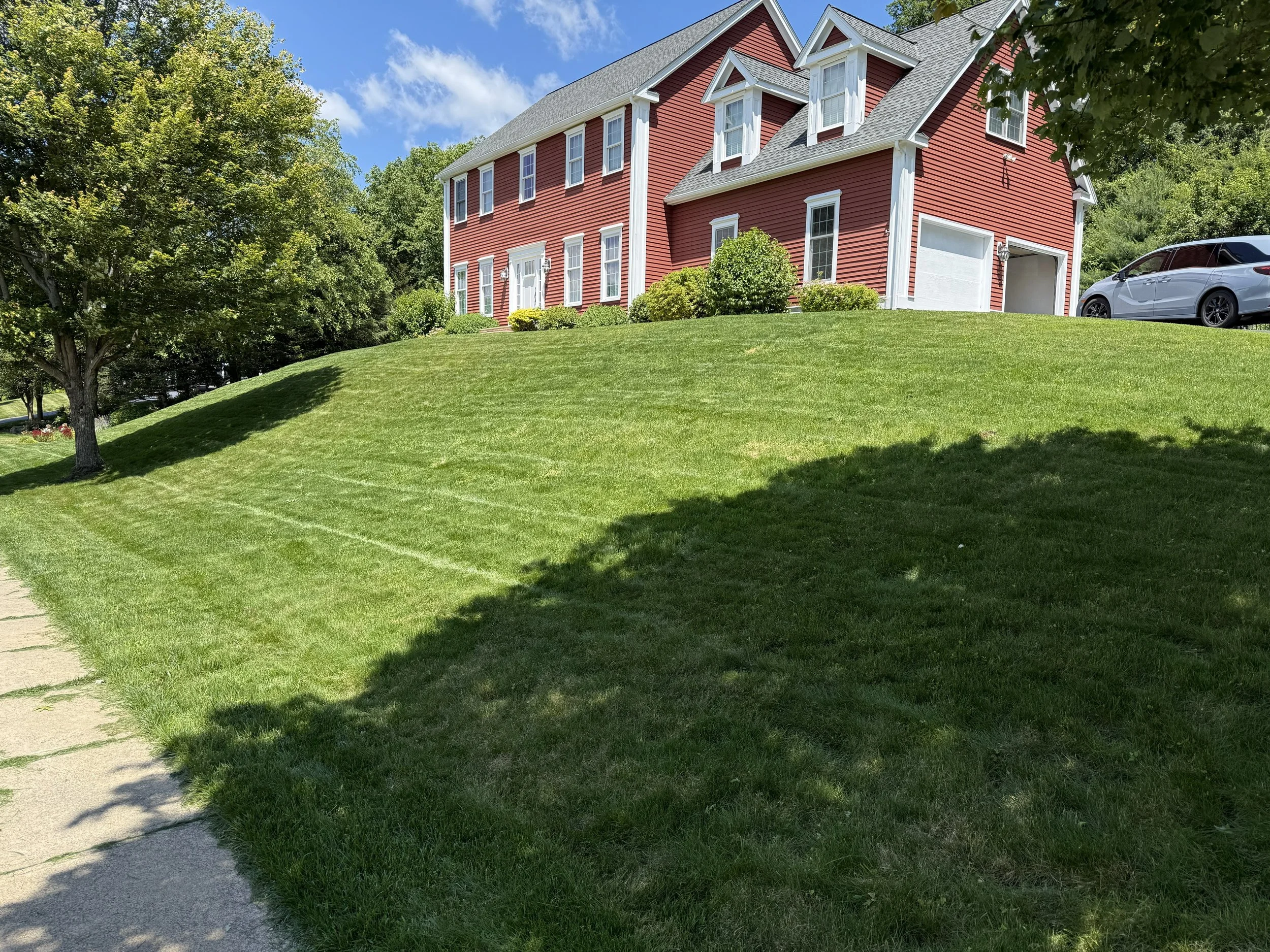 A red multi-story house with white trim, a garage, and a driveway, situated on a lush green lawn with a sidewalk on the left side. There are trees and bushes around the house, and a clear blue sky with some clouds.