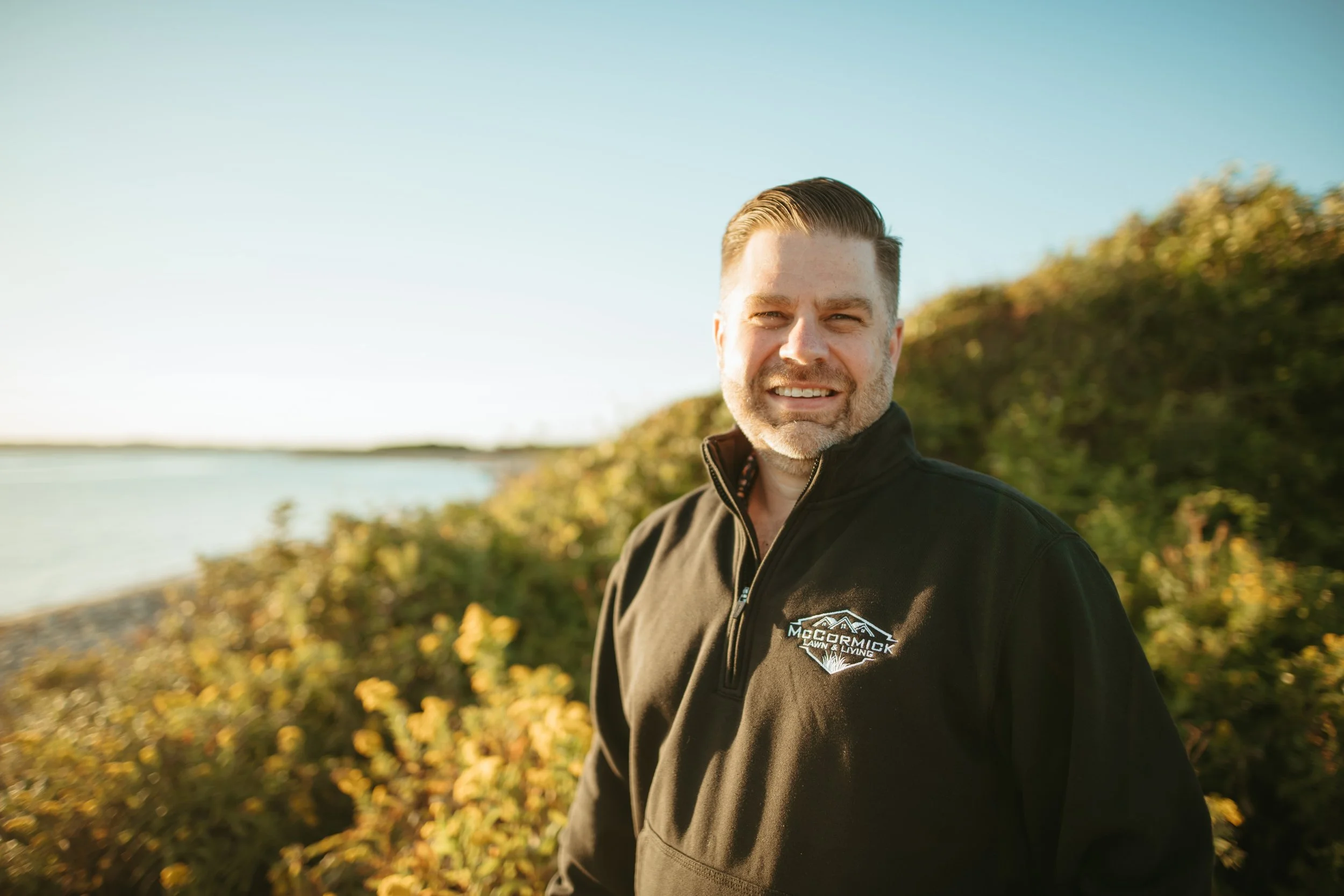 Man smiling outdoors by the water during daytime, wearing a black jacket with a logo.