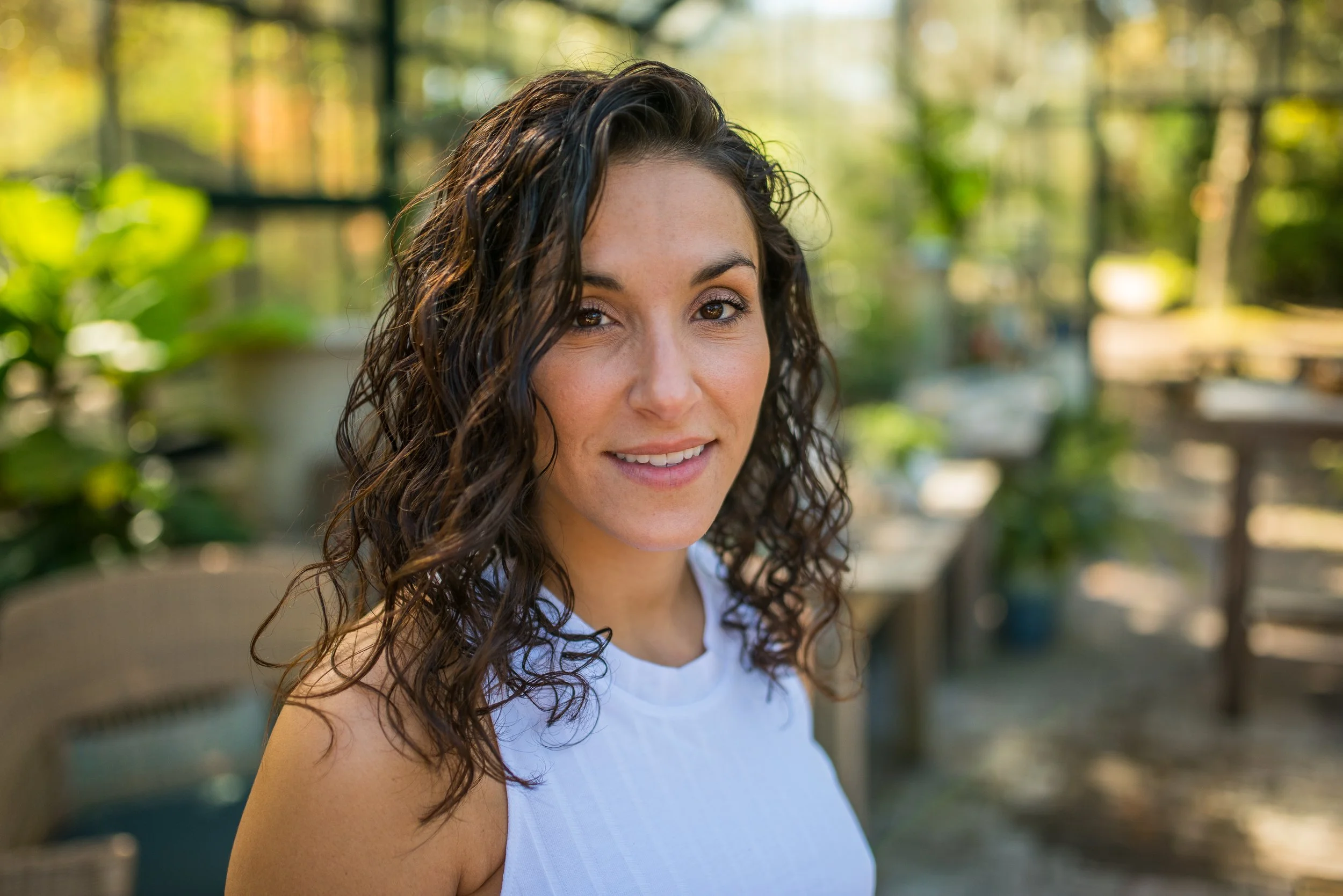 Amanda Bruno, A woman with curly dark hair outdoors, smiling, in a natural setting with trees and sunlight in the background.
