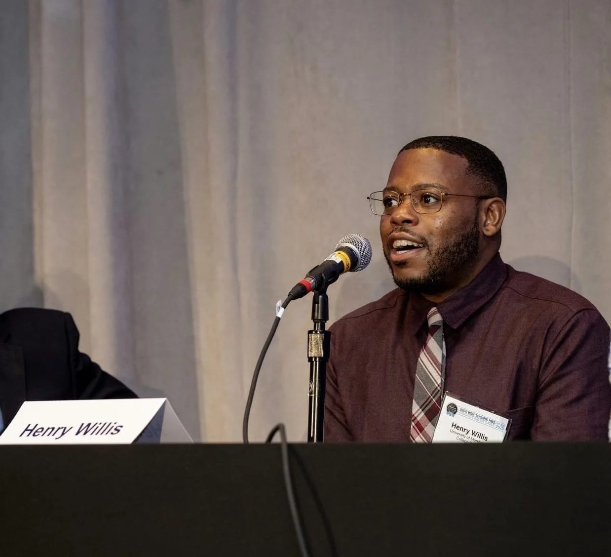 A man speaking into a microphone at a panel discussion, wearing glasses, a brown shirt, and a striped tie. A name card on the table reads "Henry Willis."