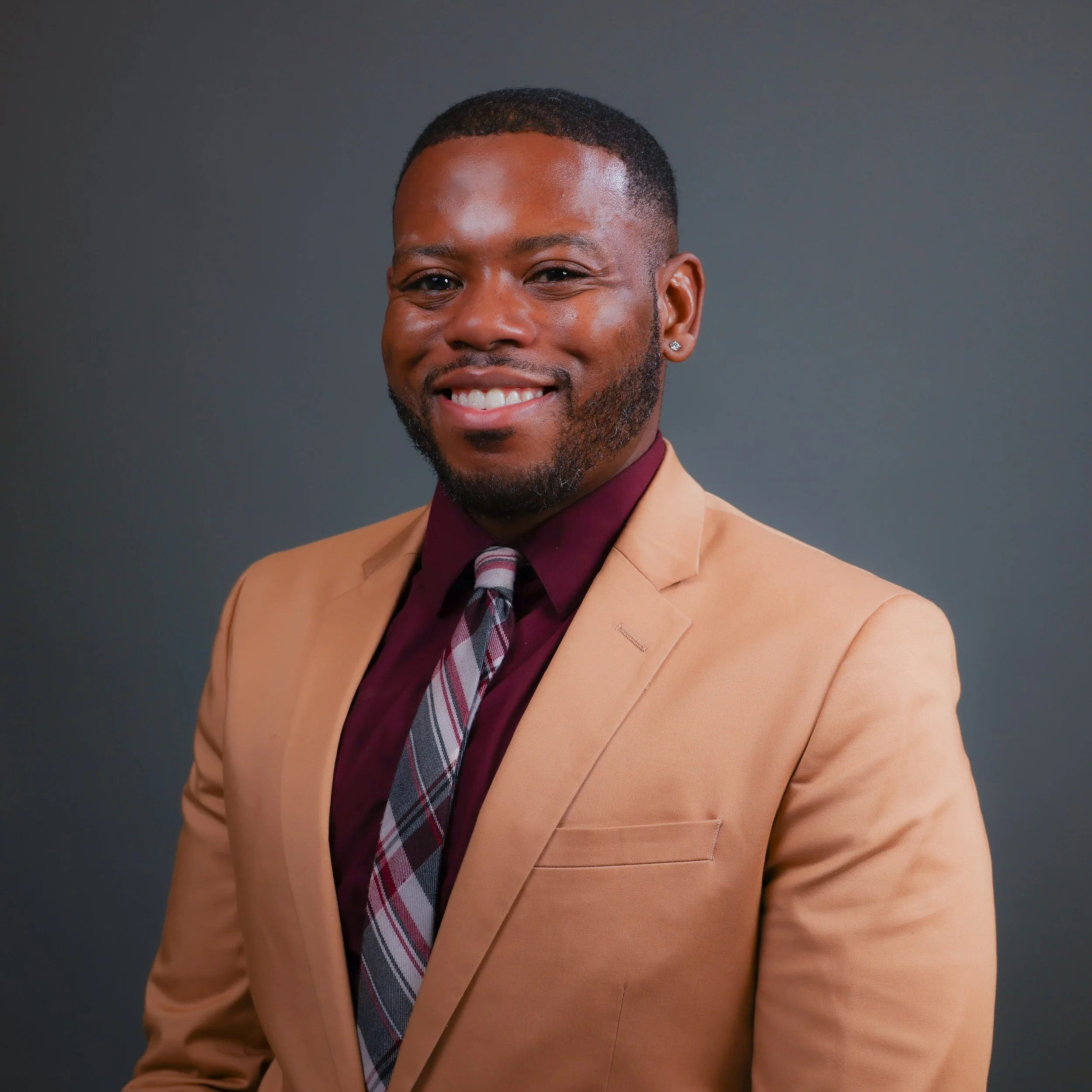 Man in a tan suit with a burgundy shirt and plaid tie smiling against a gray background.