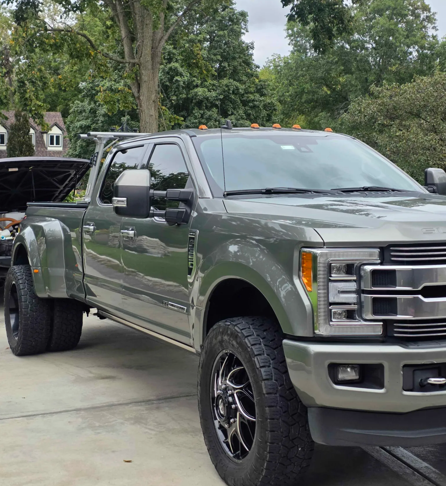 A silver pickup truck with an open hood parked on a concrete driveway in front of green trees and a house with a sloped roof.