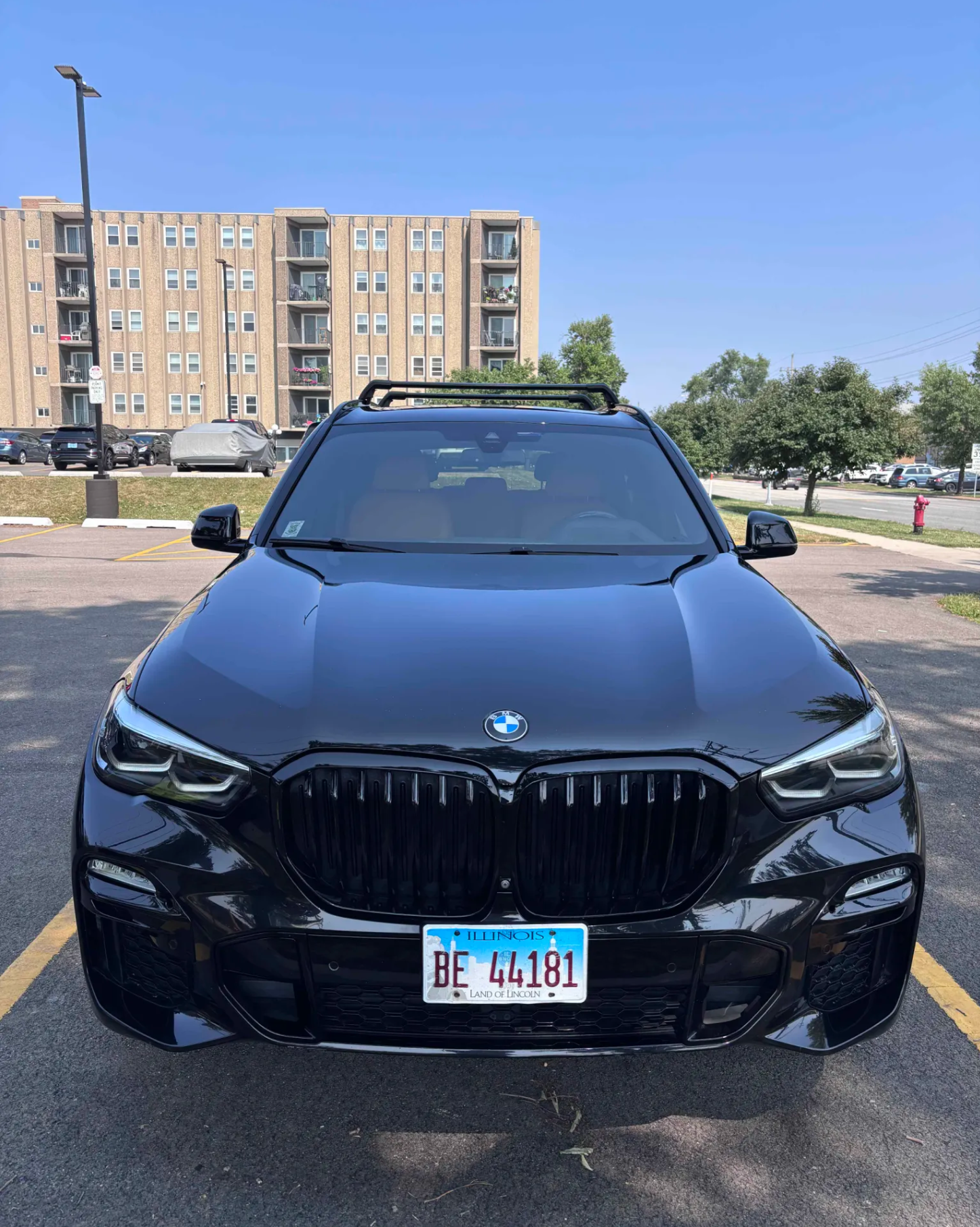 Black BMW car with Illinois license plate 'BE 44181' parked in a lot with a residential building and green trees in the background under a clear blue sky.