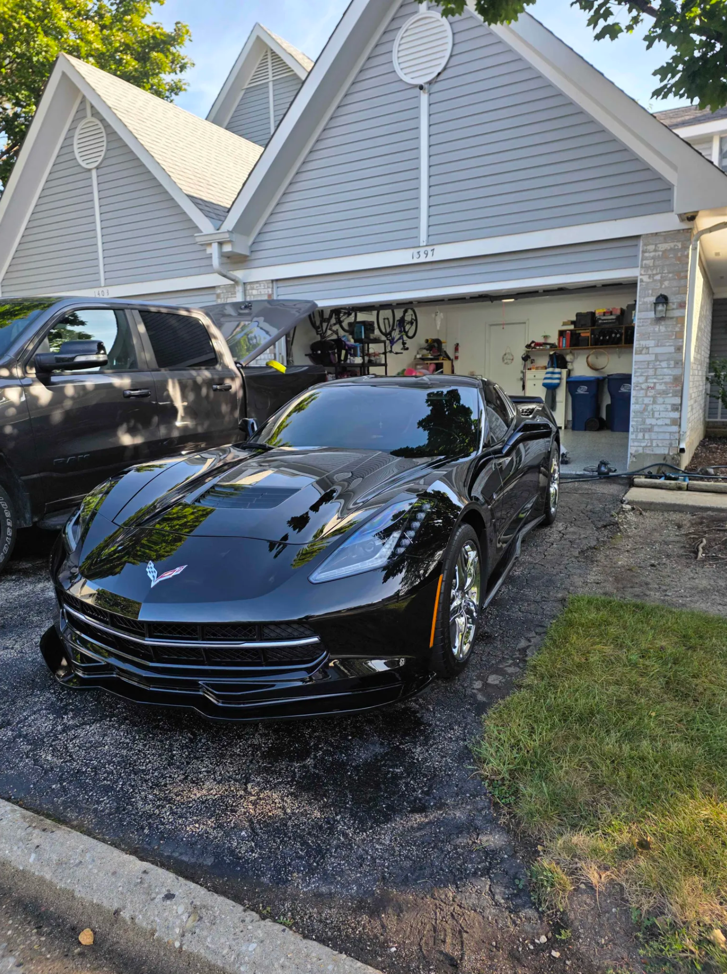 Black sports car with a Chevrolet logo parked in front of a garage, with a black pickup truck and garage tools inside, in a driveway next to a house with gray siding and a gabled roof.