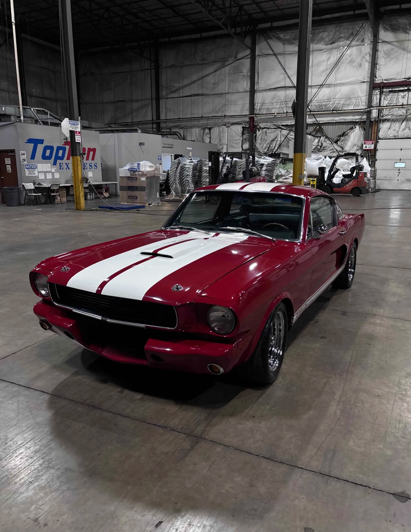 A red vintage Ford Mustang with white racing stripes parked inside a warehouse.