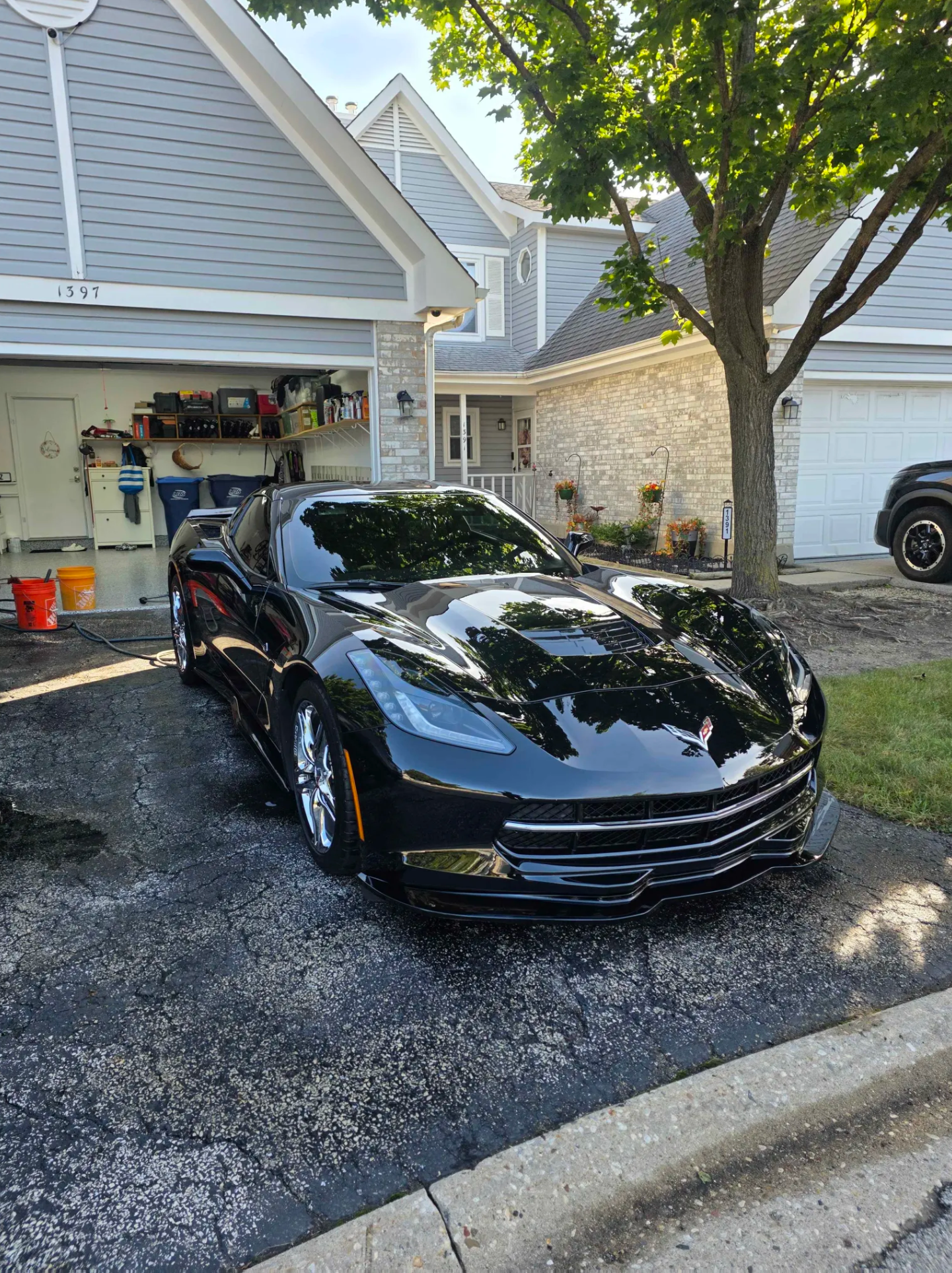 A shiny black sports car parked in the driveway of a suburban house with a garage, green lawn, and trees, on a clear day.