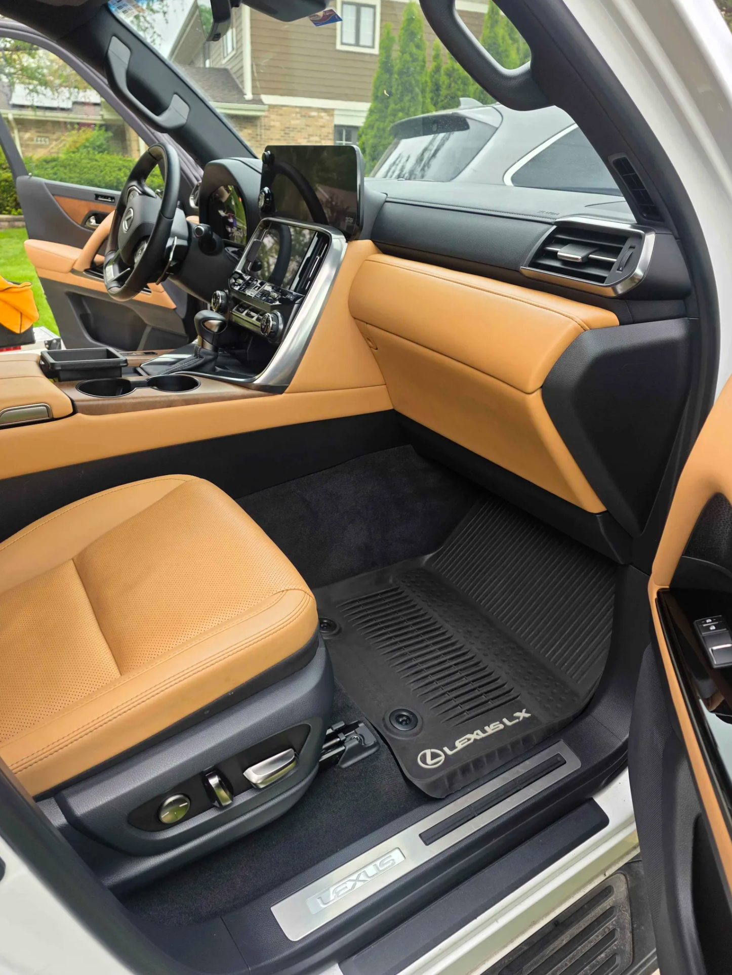 Interior of a luxury Lexus vehicle showing tan leather seats, a black dashboard, and a steering wheel with a large central display screen and controls, with a black floor mat and Lexus branding visible on the door sill.
