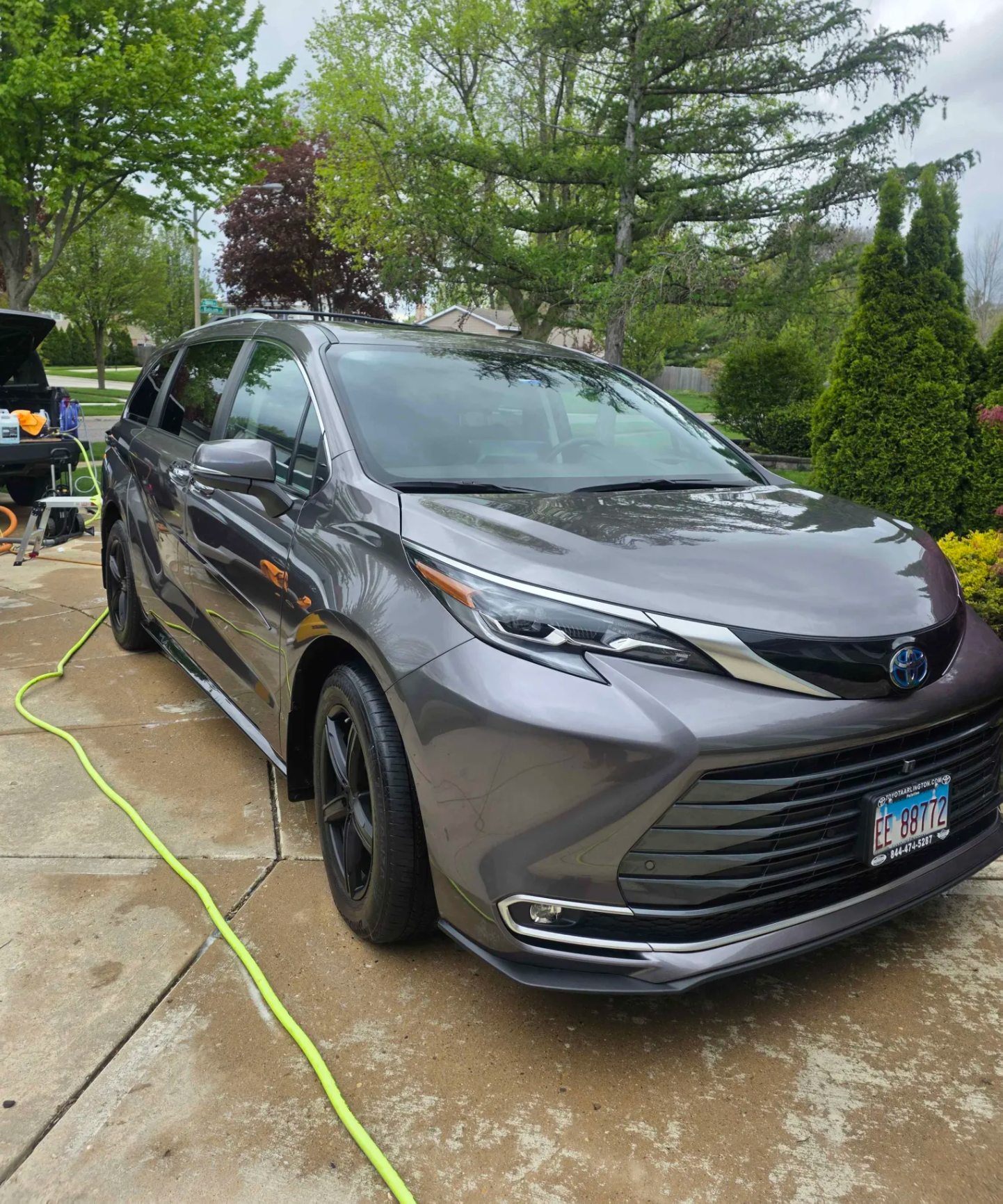 A gray Toyota electric vehicle parked in a driveway with a green garden in the background. The car is connected to a charging station, with a yellow extension cord on the wet concrete.