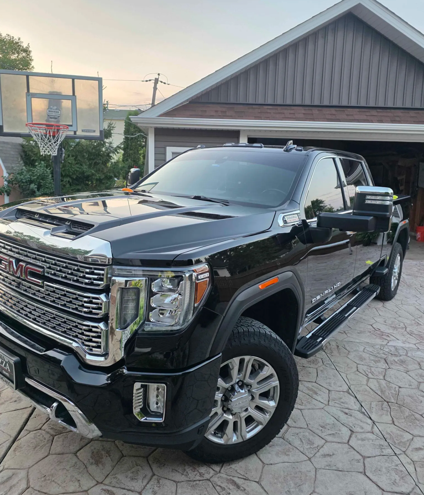 A black GMC Denali pickup truck parked in a driveway in front of a garage. In the background, there is a basketball hoop and a house with a sloped roof, trees, and electrical wires.