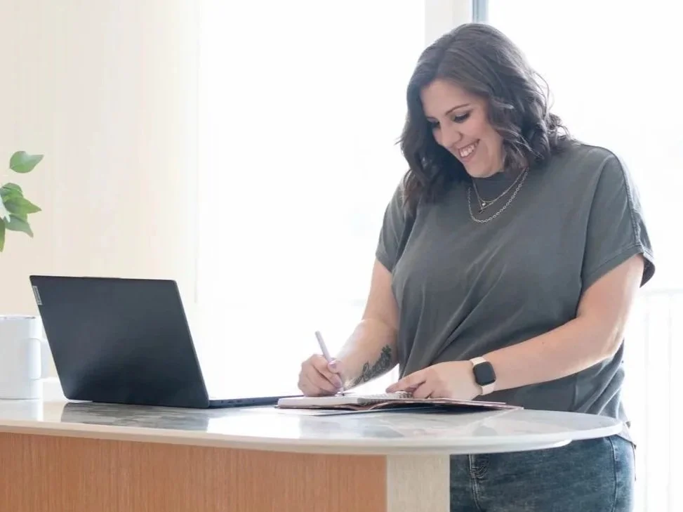 A woman with dark wavy hair, wearing a gray t-shirt and jeans, is smiling and writing in a notebook at a kitchen counter. There is a black laptop open in front of her, and a white mug on the counter.