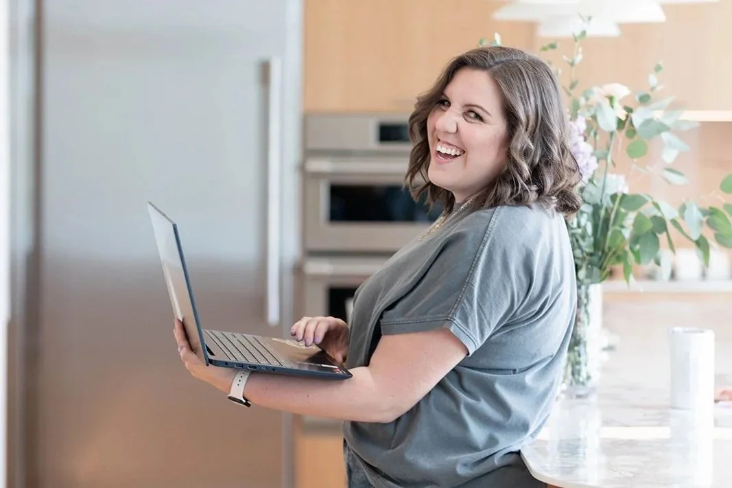 A woman with shoulder-length wavy hair smiling and holding a laptop in a bright kitchen with a plant in the background.