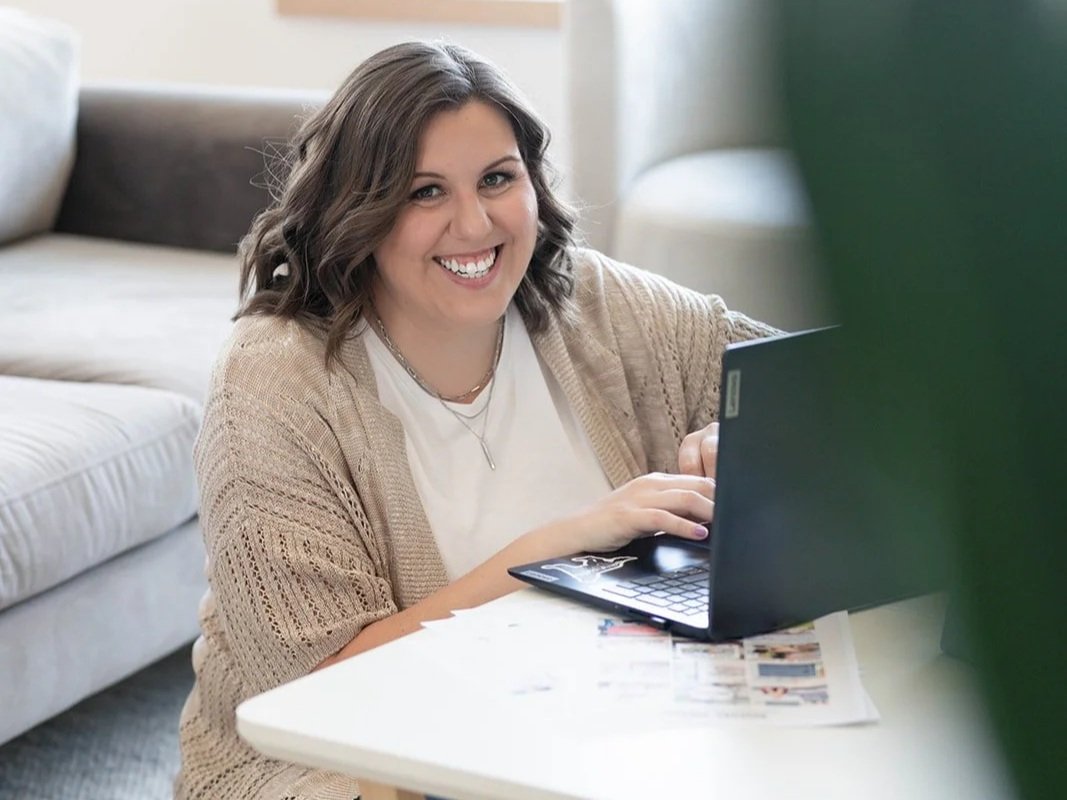 A smiling woman with shoulder-length brown hair working on a black laptop at a white table in a cozy living room.