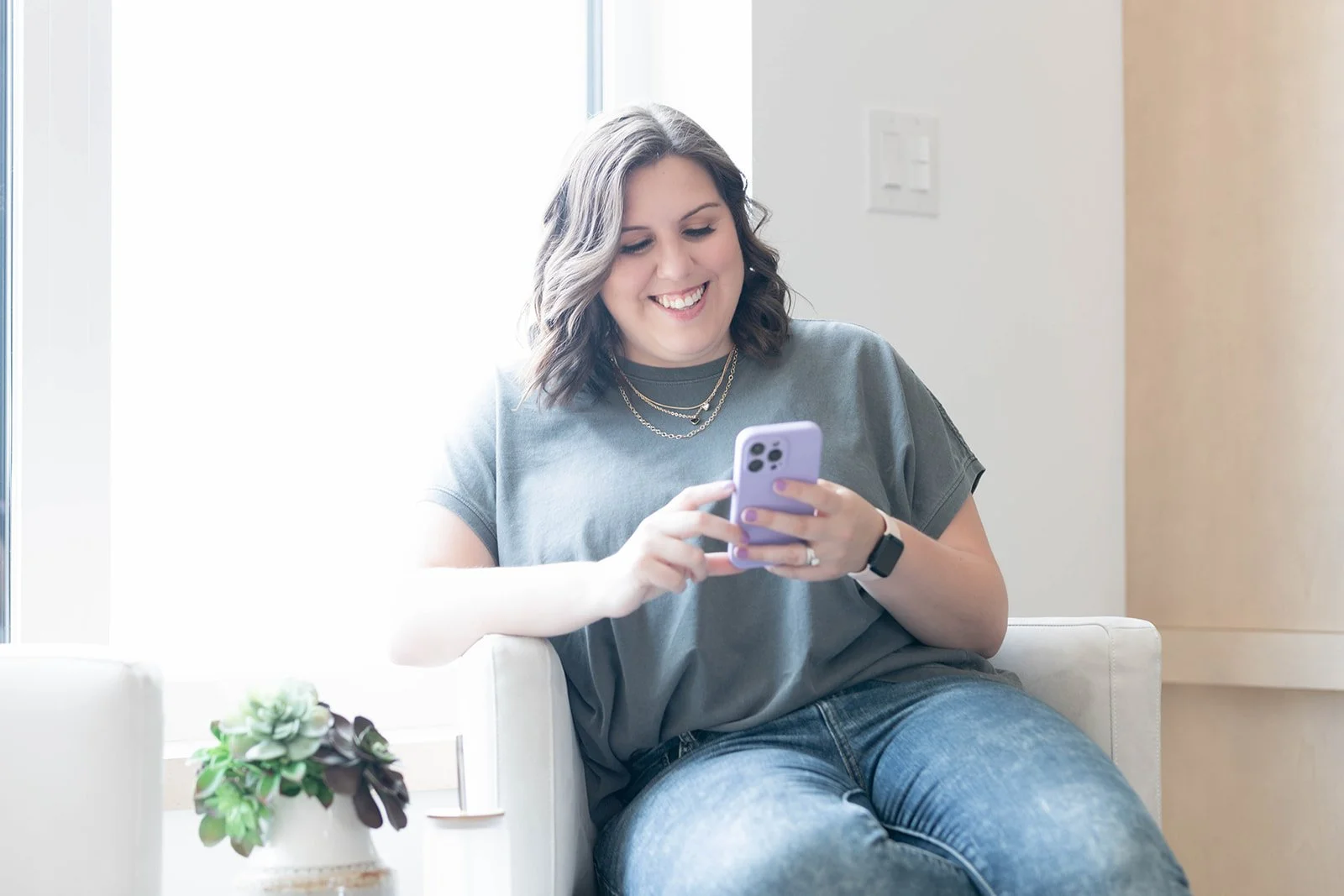 A woman with shoulder-length brown hair smiling while looking at her phone, sitting on a white chair in a bright room with a small plant on a table beside her.
