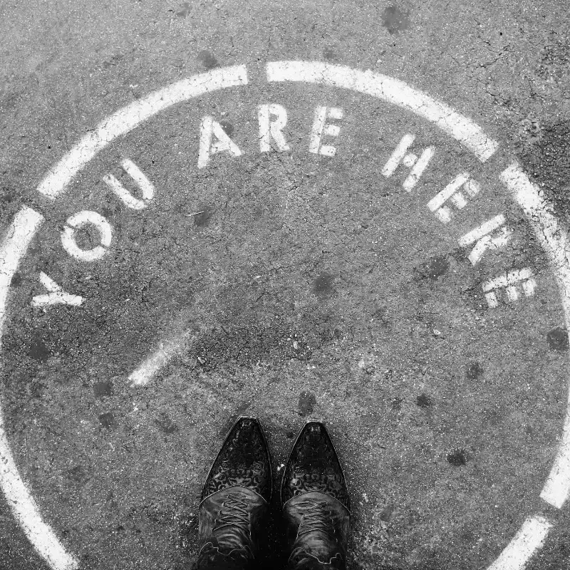 POV shot of a person looking down at their shoes on a concrete ground with the words “YOU ARE HERE” written in bold on the surface