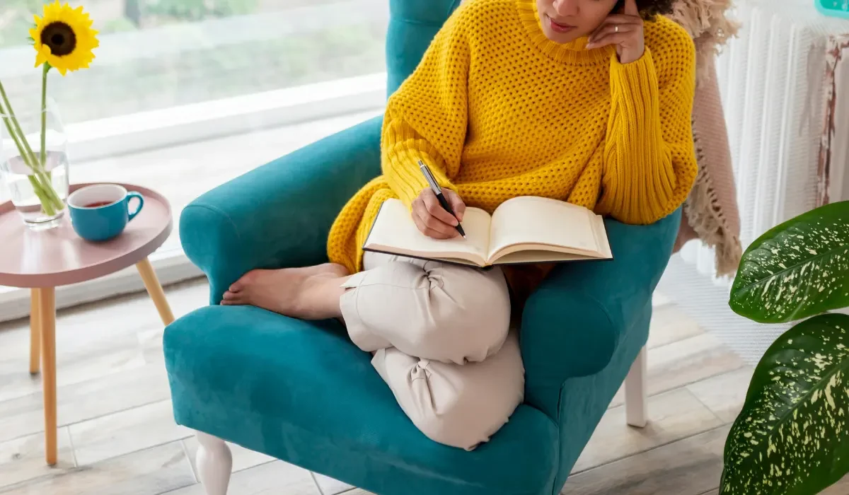 Woman in a yellow sweater writes in a notebook while seated in a blue armchair.