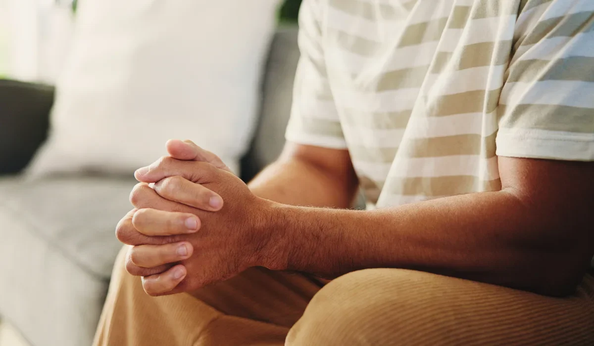 A person sits on a couch, their body slightly tense, with fingers tightly clasped together, conveying stress or anxiety.