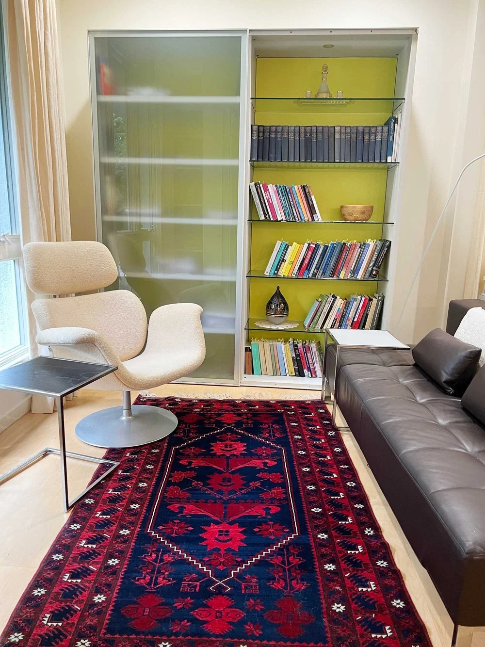 Living room with a beige swivel chair, a black sofa, a small side table, a red and black patterned rug, and a tall bookshelf with books and decorative items against a bright green accent wall.