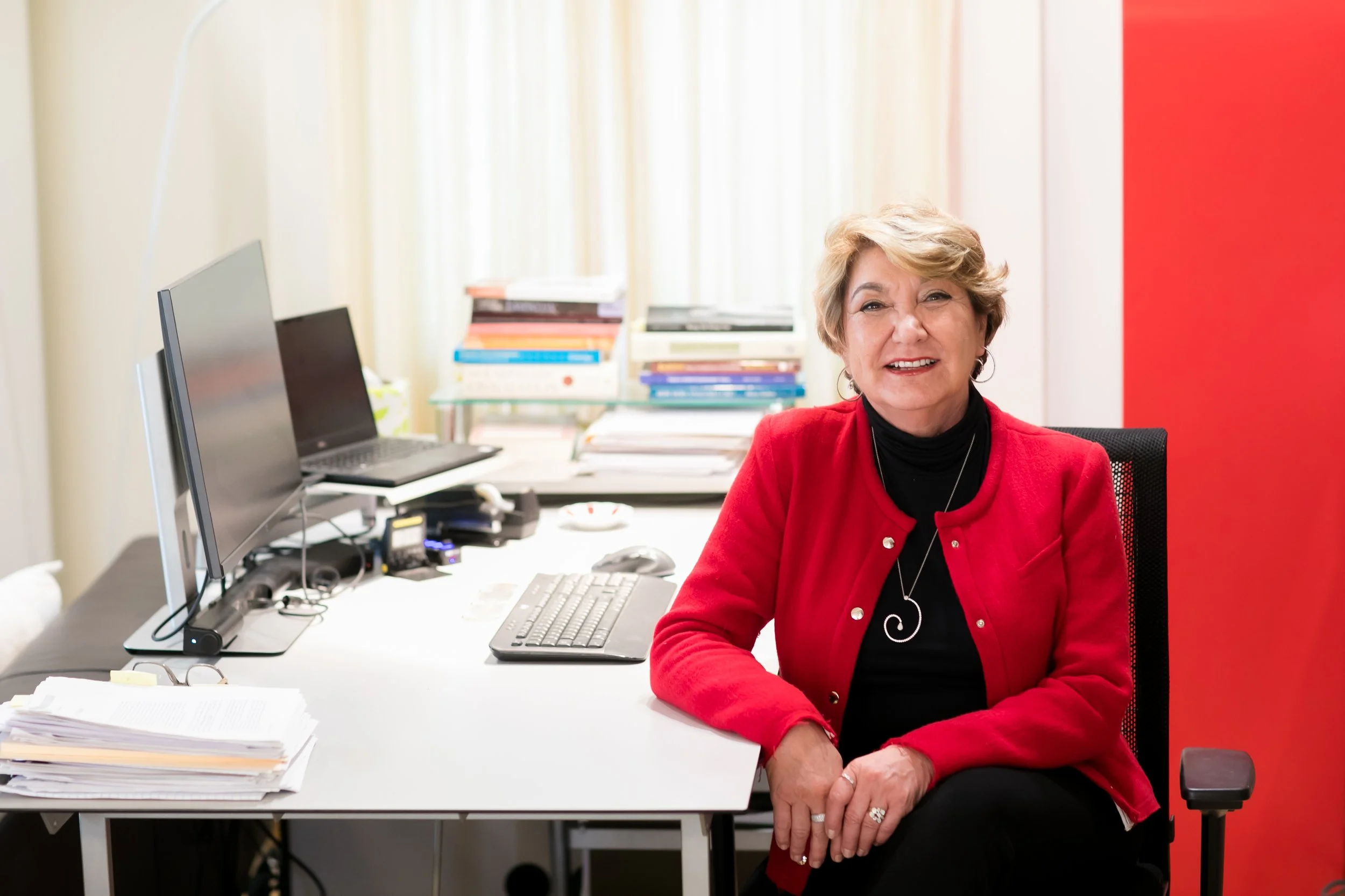 Sule Ozler sitting at a cluttered office desk with two computer monitors, stacks of books and papers, and a keyboard, in a brightly lit room with a red wall on the right.