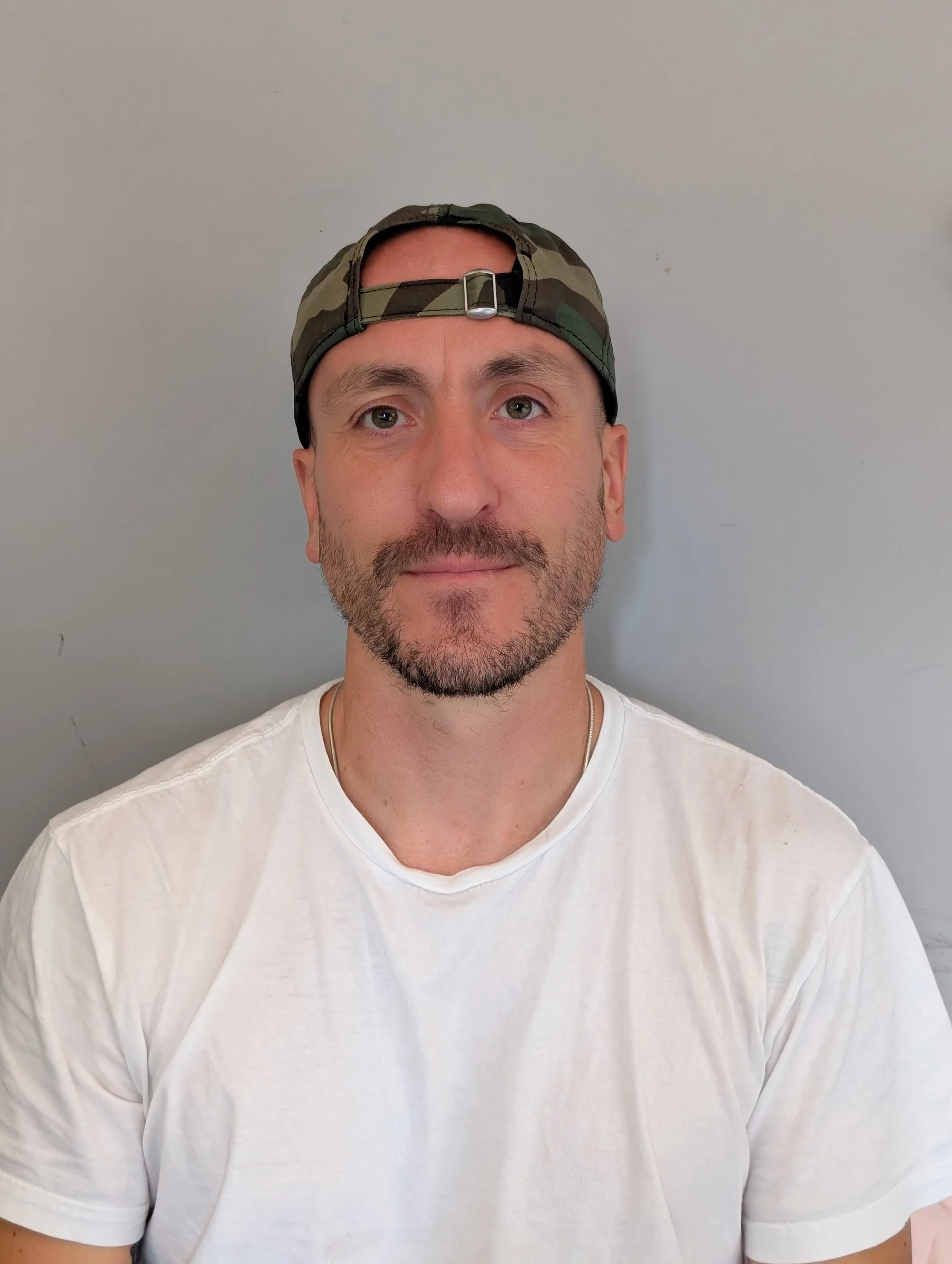 A man wearing a white t-shirt and a camouflage cap with a back strap. He has short hair, a beard, and is looking at the camera against a plain gray background.