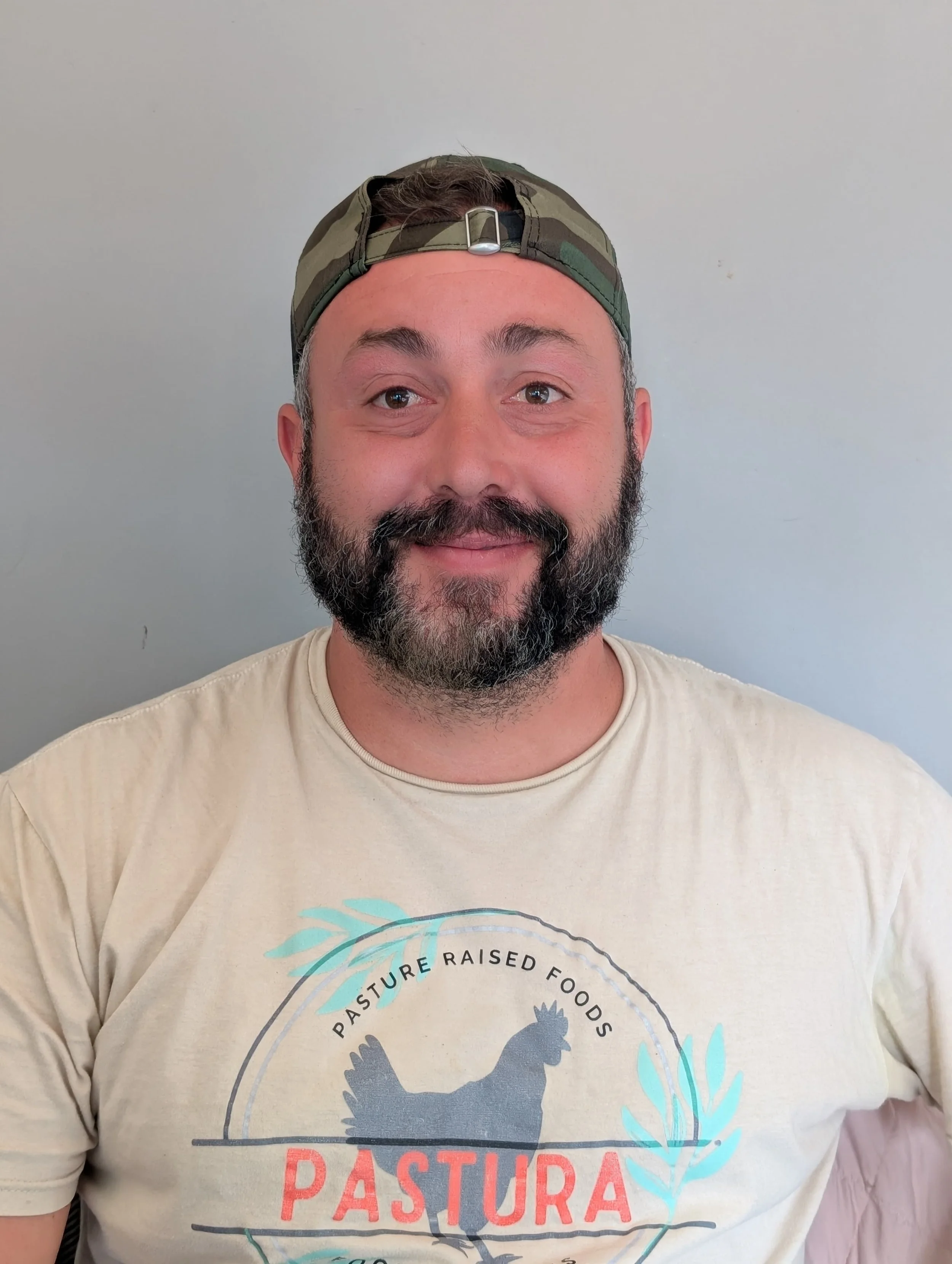 A man with a beard and mustache, wearing a camouflage baseball cap backward and a beige T-shirt with a print of a chicken and the words "Pasture Raised Foods" and "Pastura" on it.