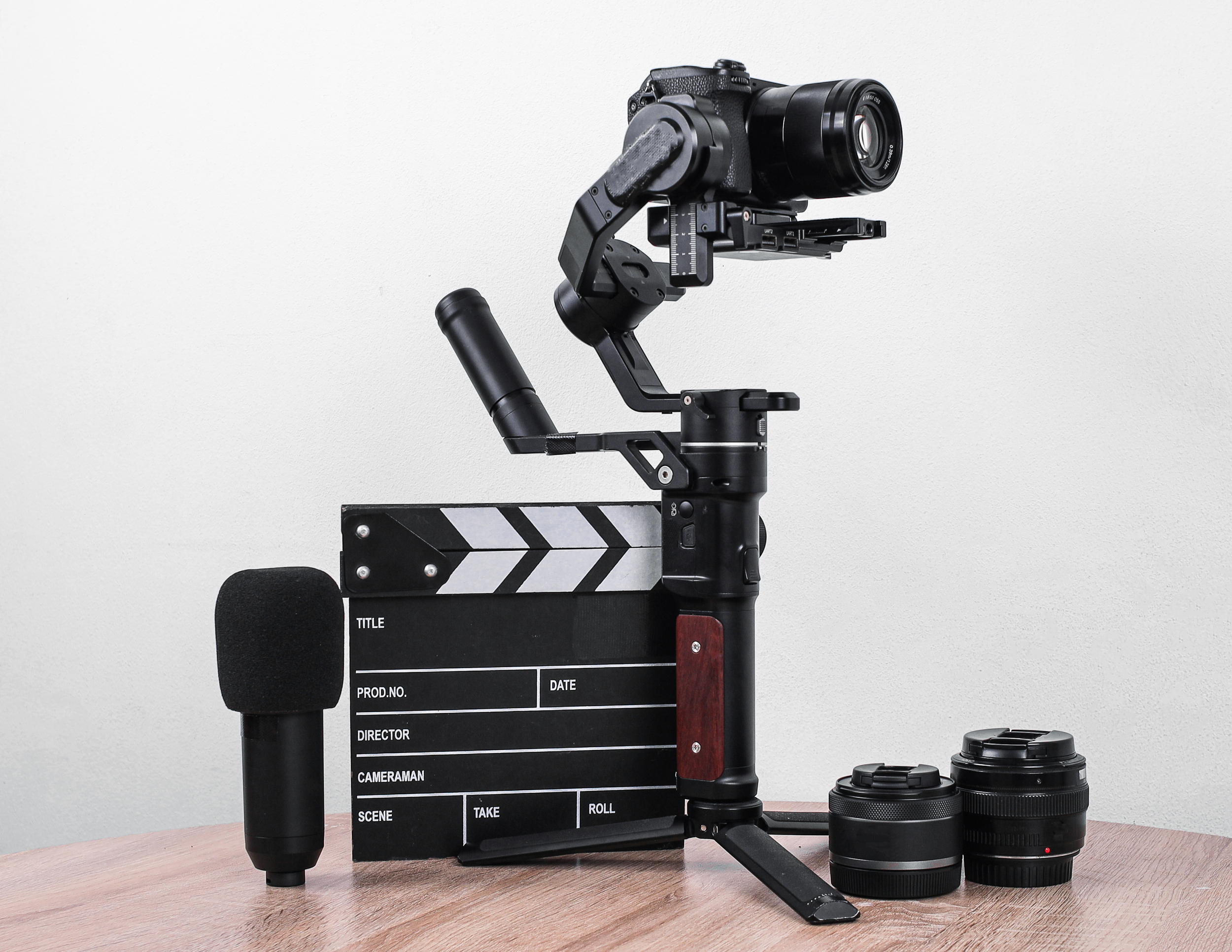 Camera stabilizer rig with mounted camera, two camera lenses, a microphone, and a film slate on a wooden table.