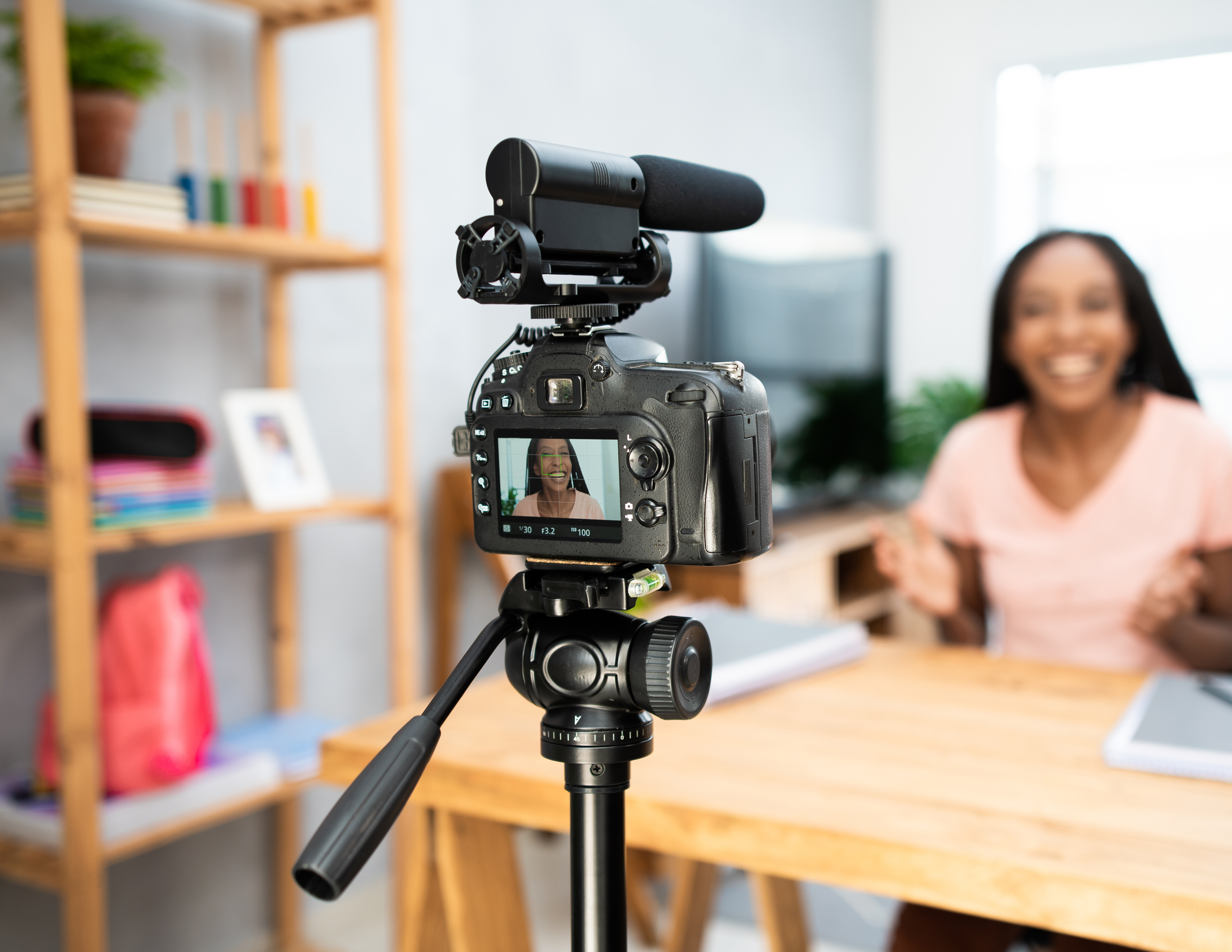 A woman is being recorded on a DSLR camera with a microphone attached, capturing her mid-sentence with her smiling face reflected on the camera screen. The background shows a wooden bookshelf, a desk, and decorative items.
