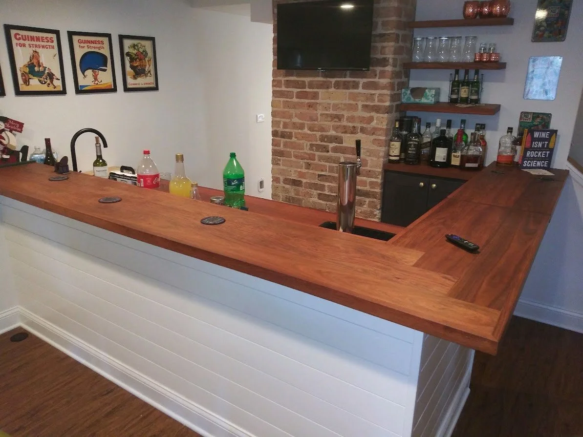 Home bar with wooden counter, liquor bottles on shelves, TV on brick wall, decorative signs, and various drinks on the counter.