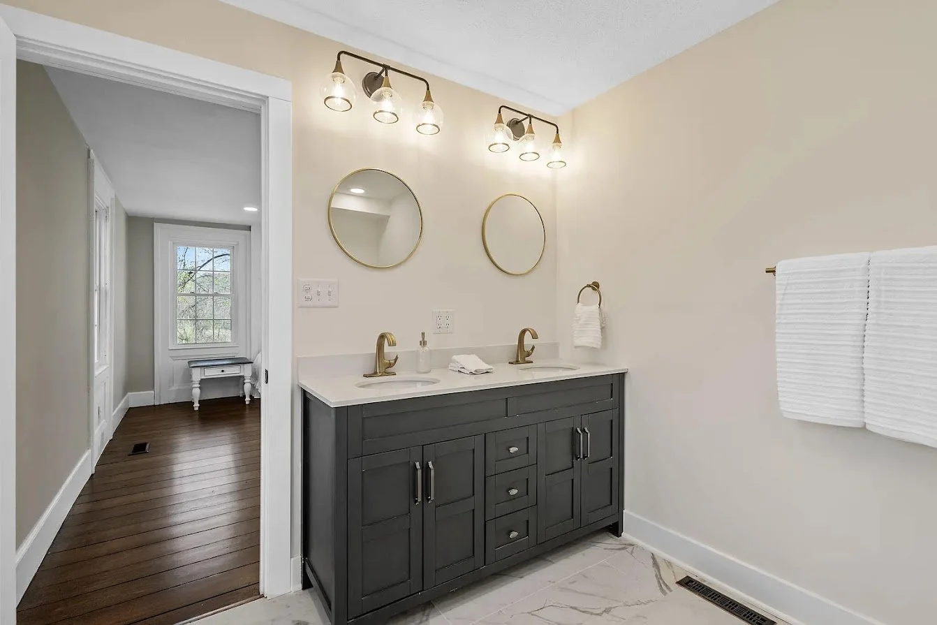 Modern bathroom with double sink vanity, dark gray cabinetry, two round mirrors, and gold fixtures. There are four light bulbs fixture above the mirrors, a towel hanging on a ring, and a window visible in the adjacent room.
