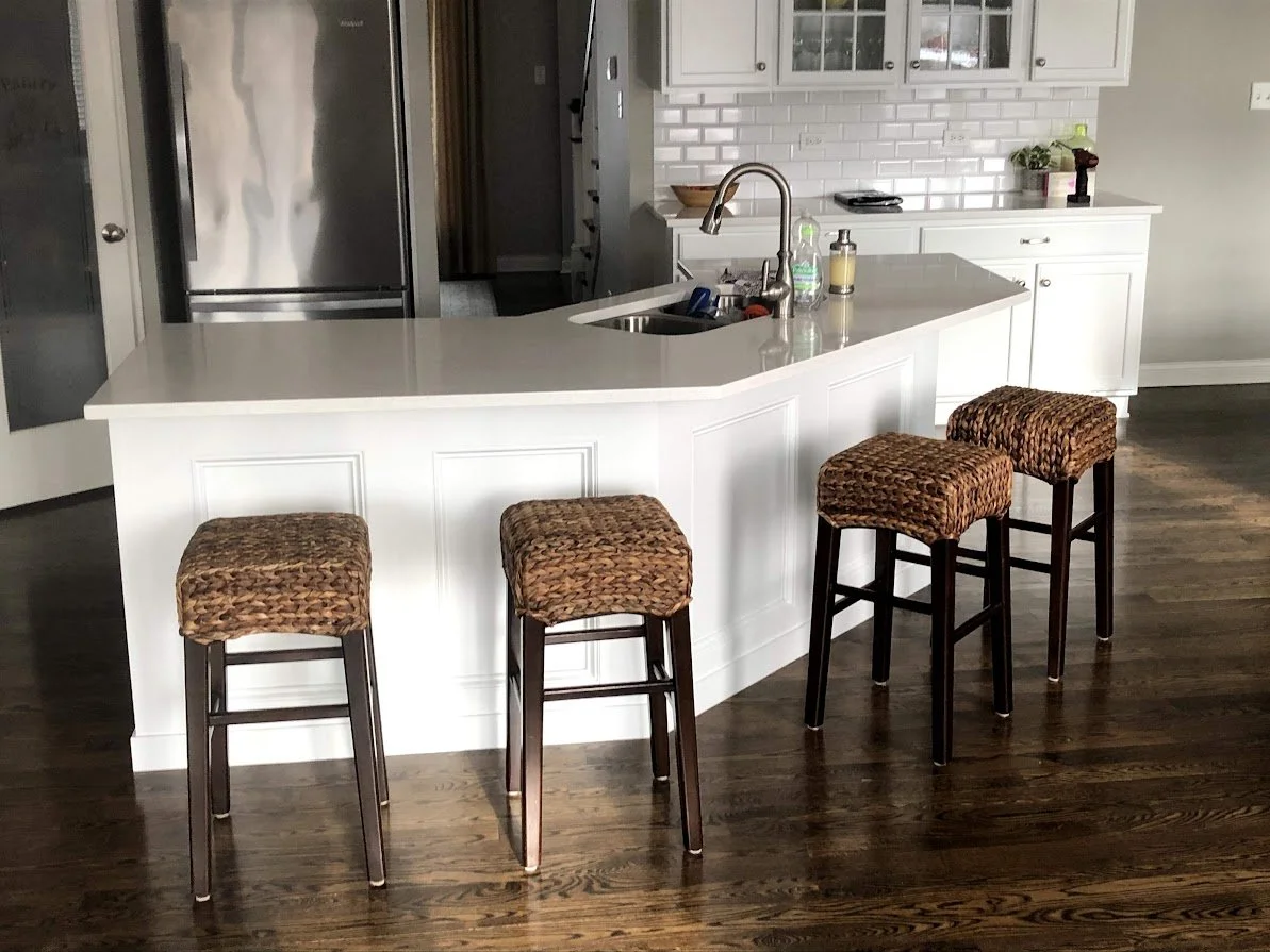 Modern kitchen with white island counter, four bar stools with woven seats, stainless steel refrigerator, white cabinetry, and dark hardwood floors.